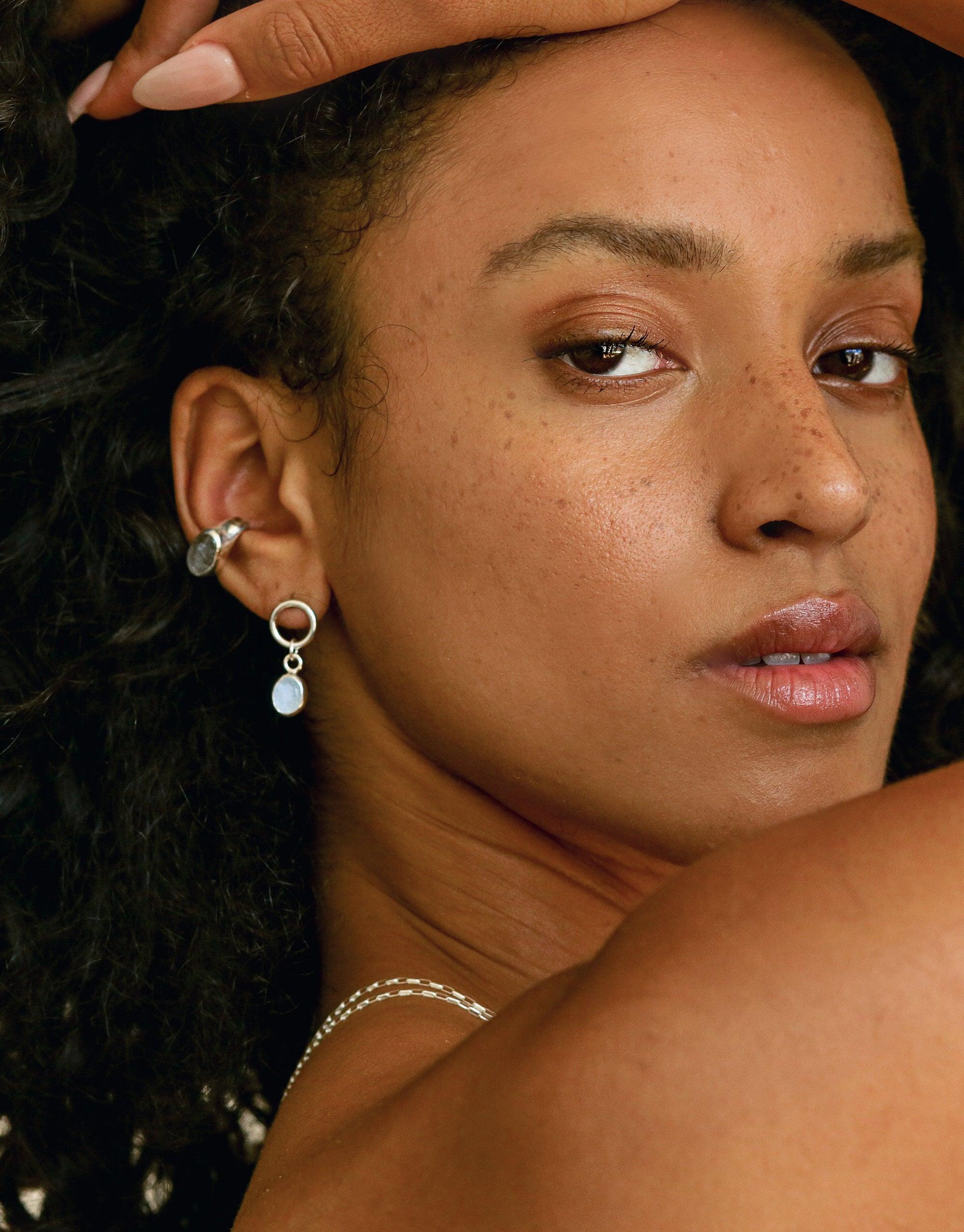 Close-up of model wearing Oval Dangling Earrings, featuring delicate silver hoops with translucent oval charms. Earrings complement natural beauty, highlighting freckles and curly hair. Handcrafted jewelry showcases eco-friendly design. Oval Dangling Earrings by CLED