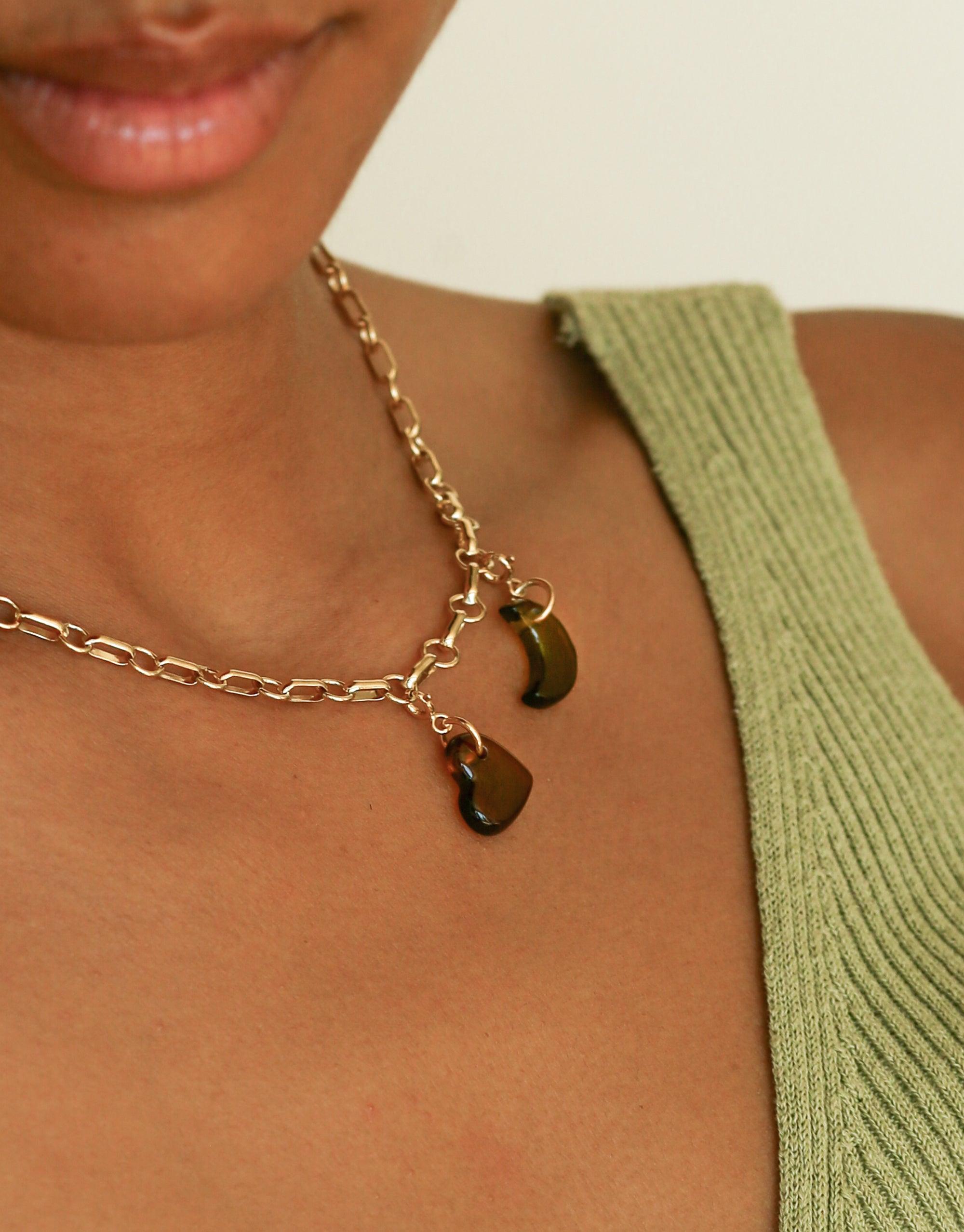 Close-up of gold chain necklace with two crescent moon-shaped charms, worn on a person's neck. Green ribbed fabric visible on shoulder. Elegant and minimalist jewelry design. Moon Icon Charm by CLED.