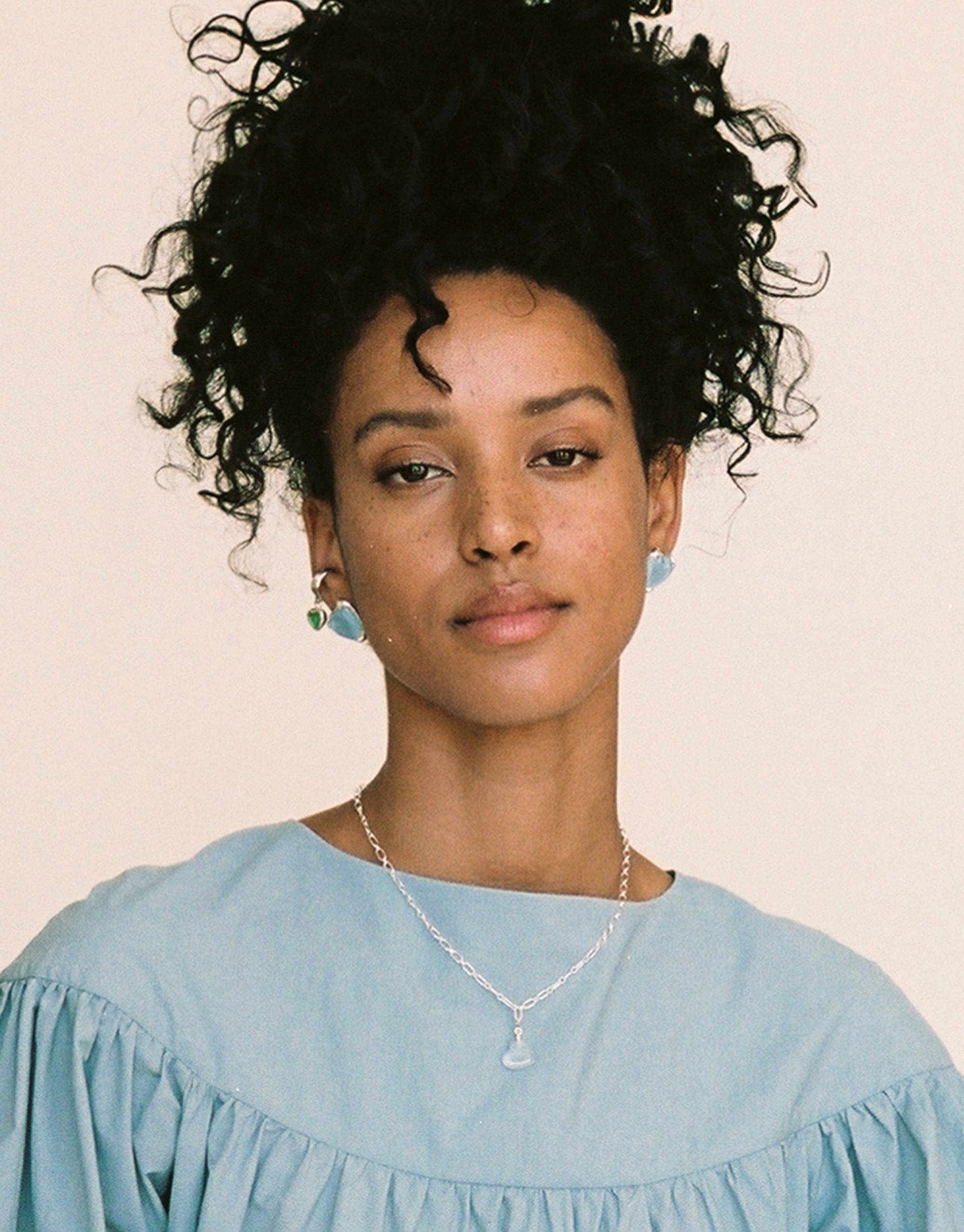 Model wearing Heart Icon Charm necklace with sterling silver chain, showcasing delicate pendant against light blue blouse. Curly hair styled in updo. Earrings visible. Elegant jewelry display for Heart Icon Charm by CLED.
