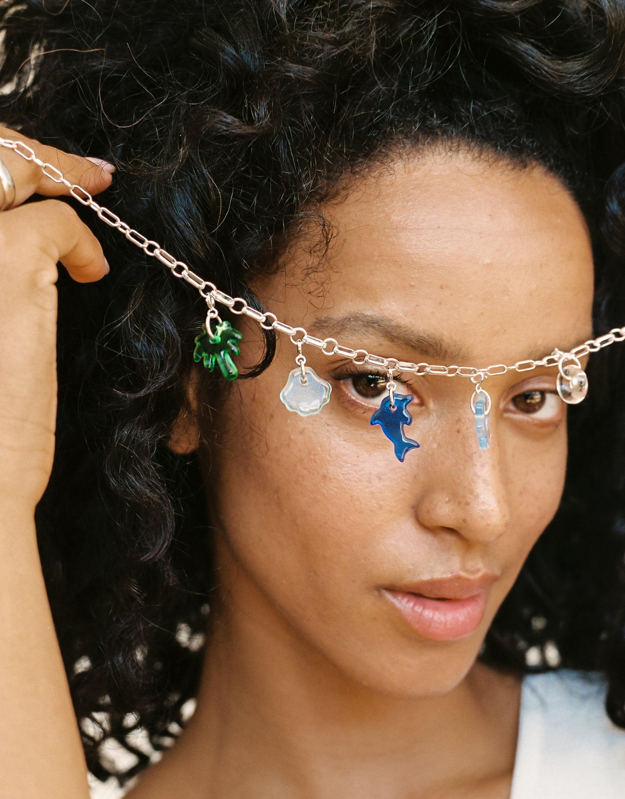 Silver chain necklace with colorful icon charms including a green shell, blue dolphin, and white cloud, worn by a person with curly dark hair. Showcases the versatility and design of the Shell | Icon Charm collection by CLED.