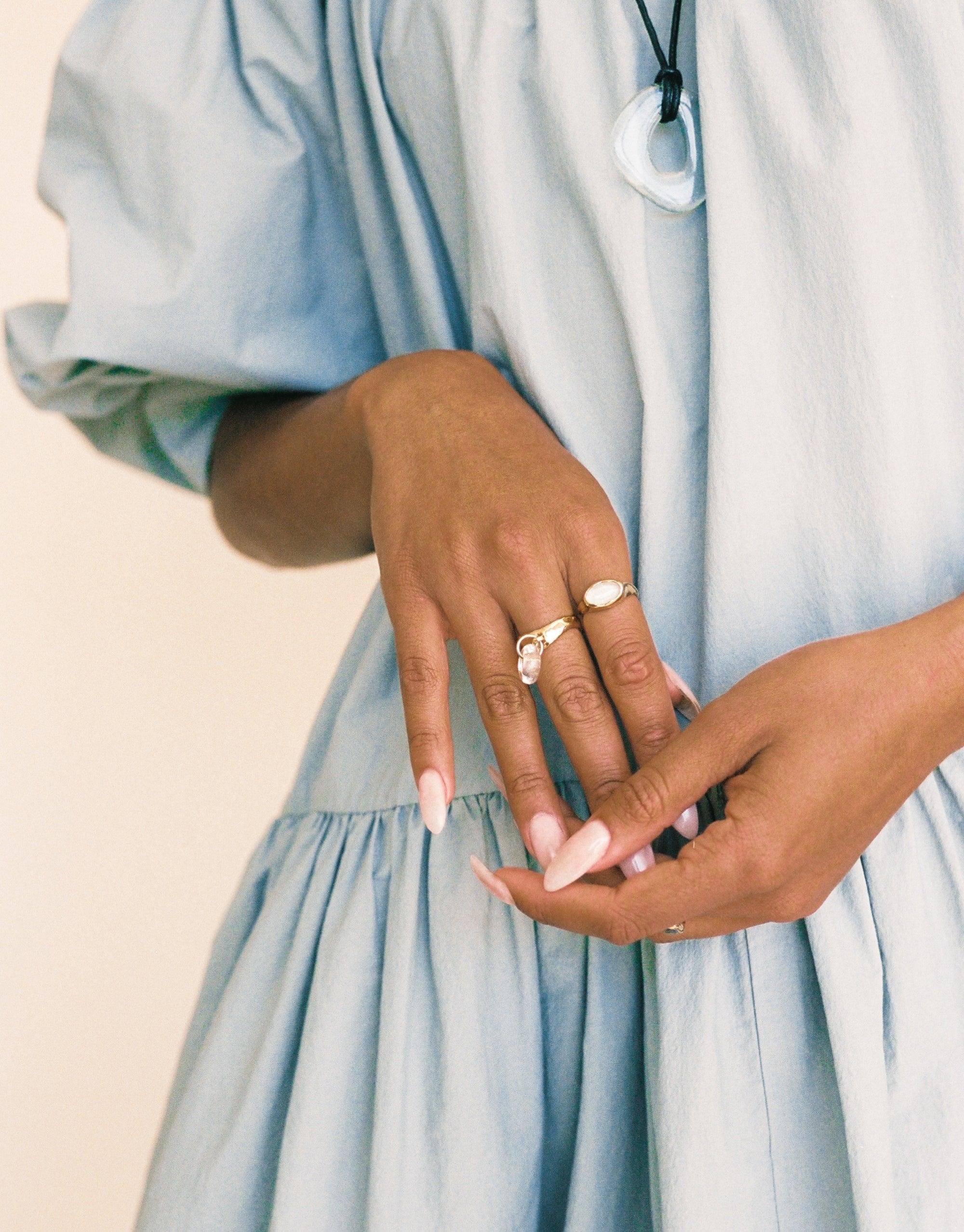 Sculptural Large Charm Necklace | A by CLED: Elegant hands wearing rings and holding a light blue dress, with a glimpse of a glass pendant necklace, showcasing the handcrafted, upcycled jewelry piece against a soft, feminine backdrop.