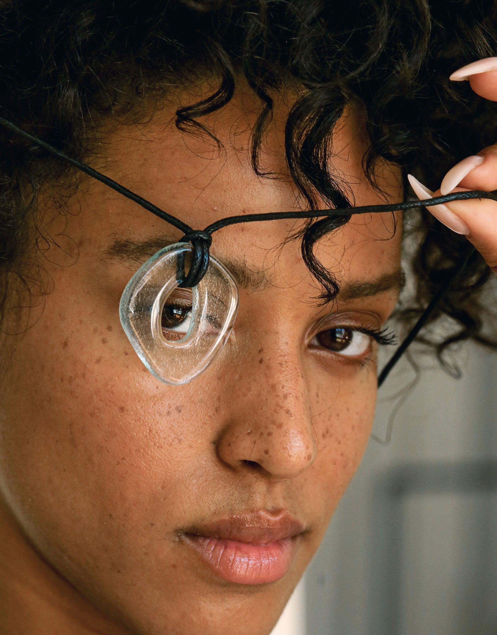 Close-up of Sculptural Large Charm Necklace featuring transparent glass pendant on black cord against curly hair and freckled skin. Sustainable upcycled jewelry design showcasing eco-friendly materials. Sculptural Large Charm Necklace | A by CLED