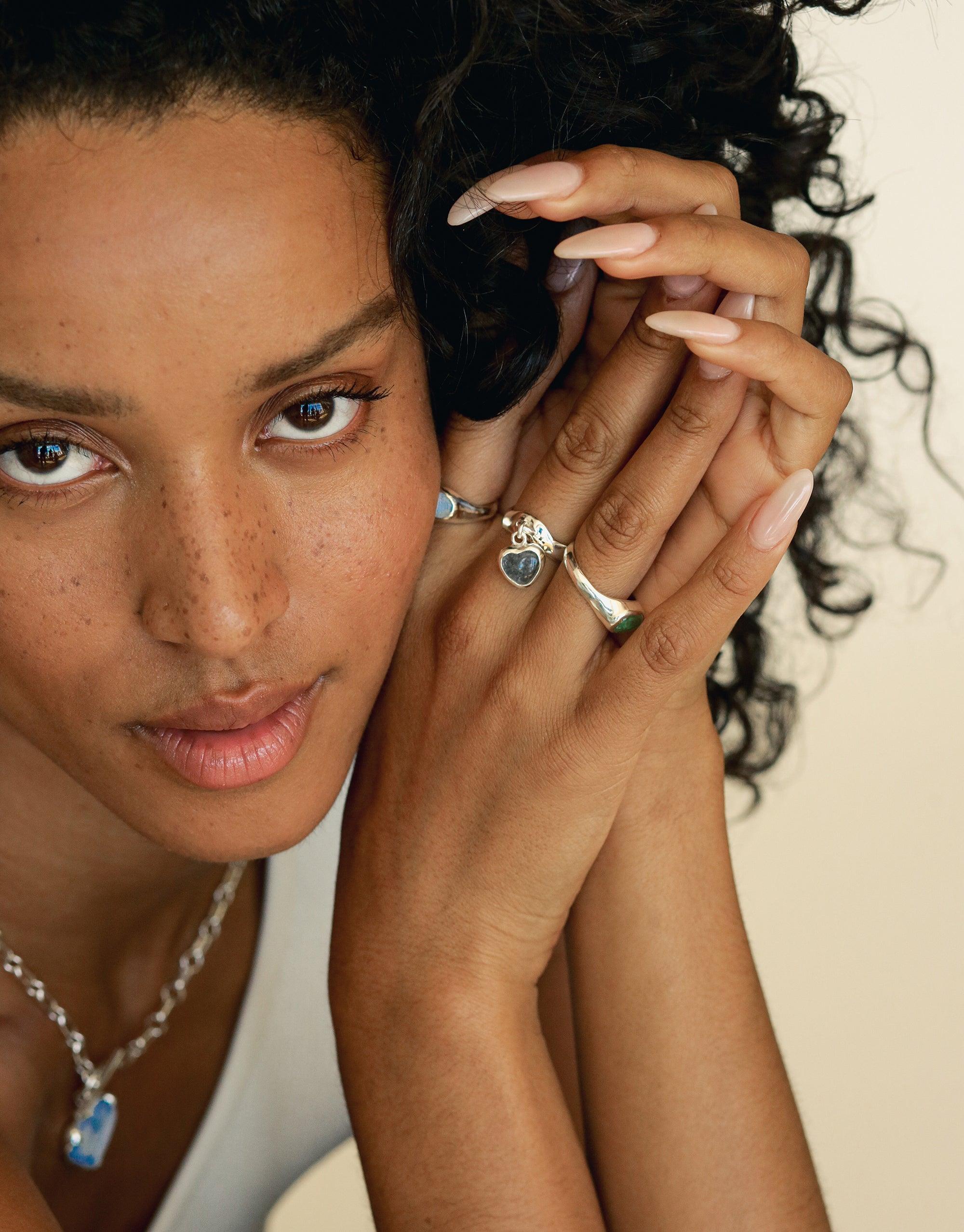 Close-up of hands wearing delicate silver rings, including the Heart Dangling Ring. Matching necklace visible. Showcases unique eco-friendly jewelry designs made from upcycled materials. Handcrafted luxury accessories by CLED.