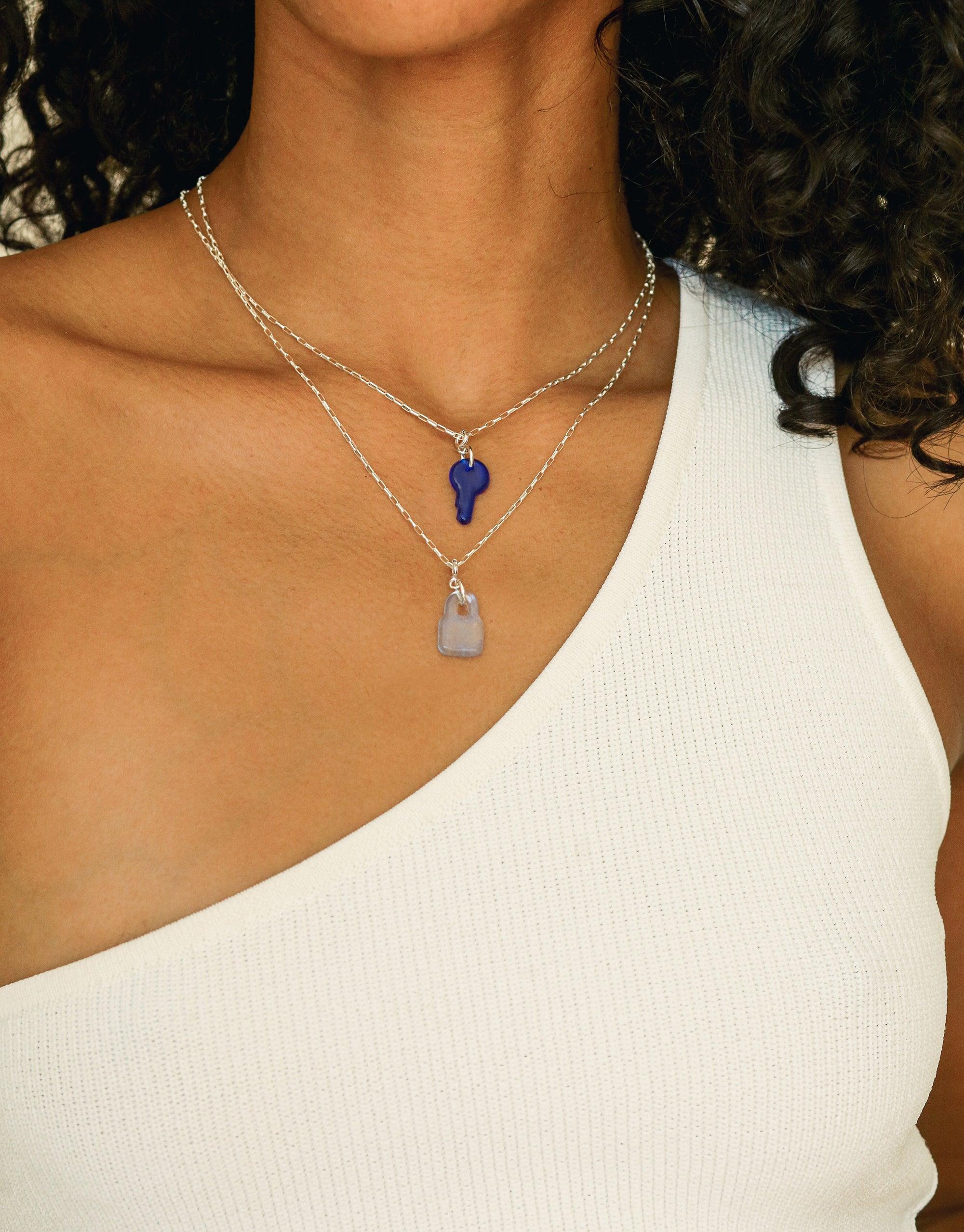 Close-up of layered silver necklaces featuring a blue key-shaped charm and a pale pink rectangular charm, worn with a white top, showcasing the versatile Key Icon Charm by CLED