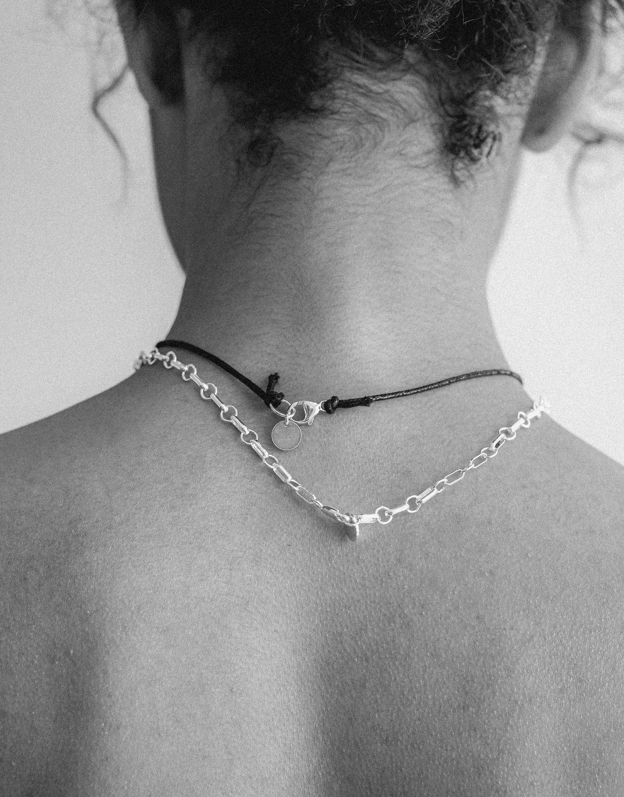 Black and white image of a person's back wearing a delicate silver chain necklace and a thin black cord necklace. The Sculptural Small Charm Necklace | C showcases elegant layering and minimalist design, highlighting CLED's commitment to sustainable, handcrafted jewelry.