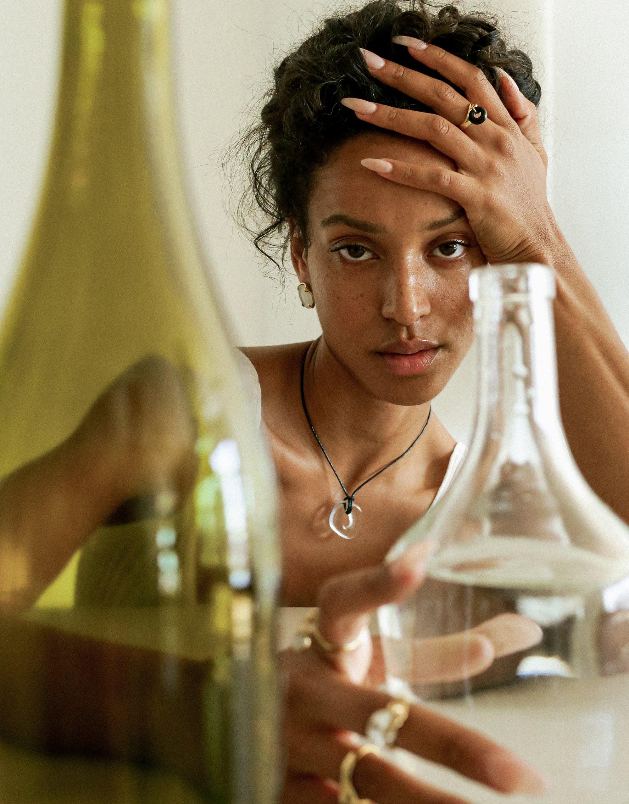 Close-up of a person wearing a delicate necklace with a small transparent charm, surrounded by glass bottles. The image showcases the Sculptural Small Charm Necklace, highlighting its subtle elegance and eco-friendly design made from repurposed glass. CLED
