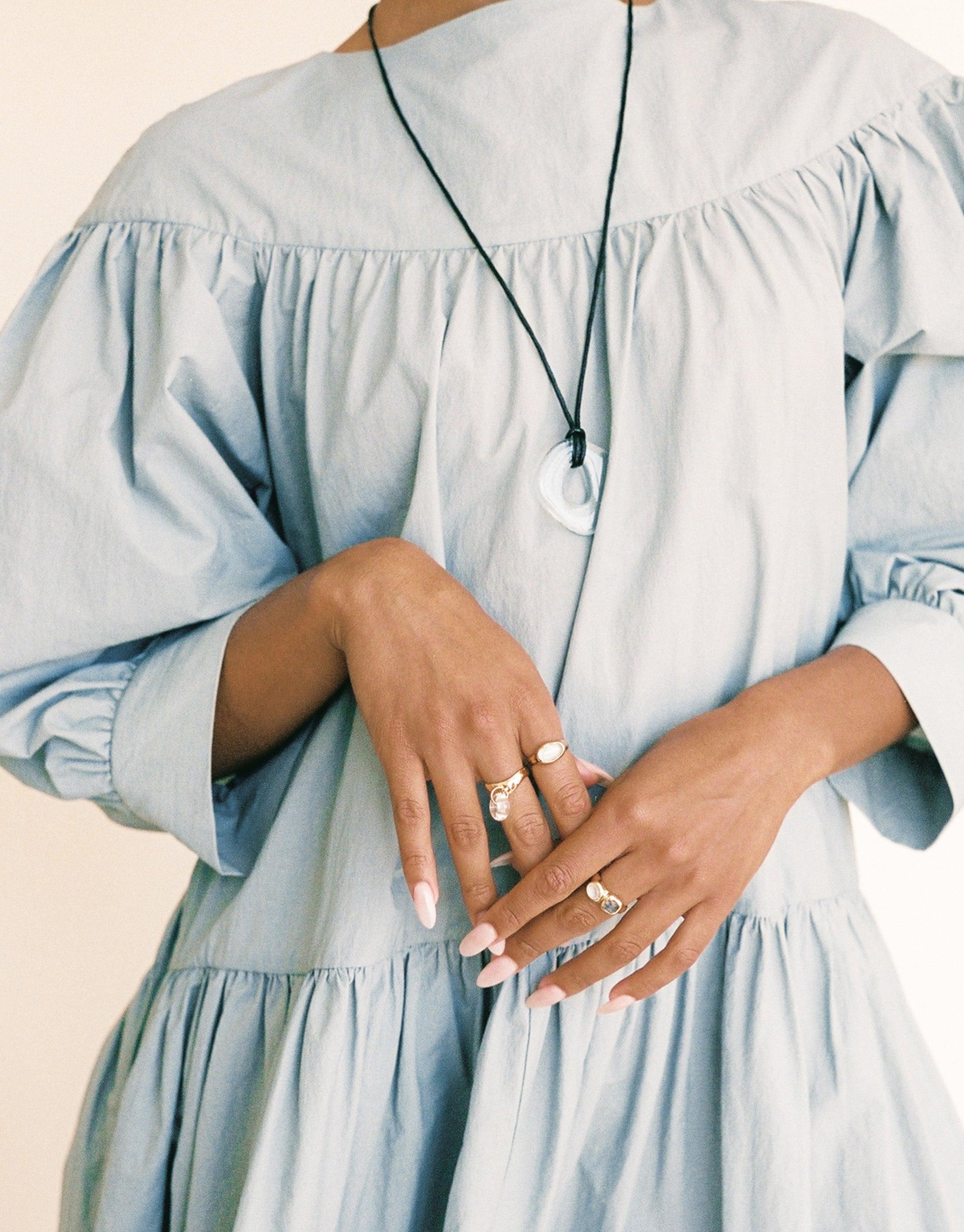 Sculptural Large Charm Necklace with white pendant on dark cord, worn over light blue ruffled dress. Hands displaying multiple rings. Eco-friendly, handmade jewelry showcasing upcycled glass charm. Adjustable length for versatile styling. Sculptural Large Charm Necklace | A by CLED.