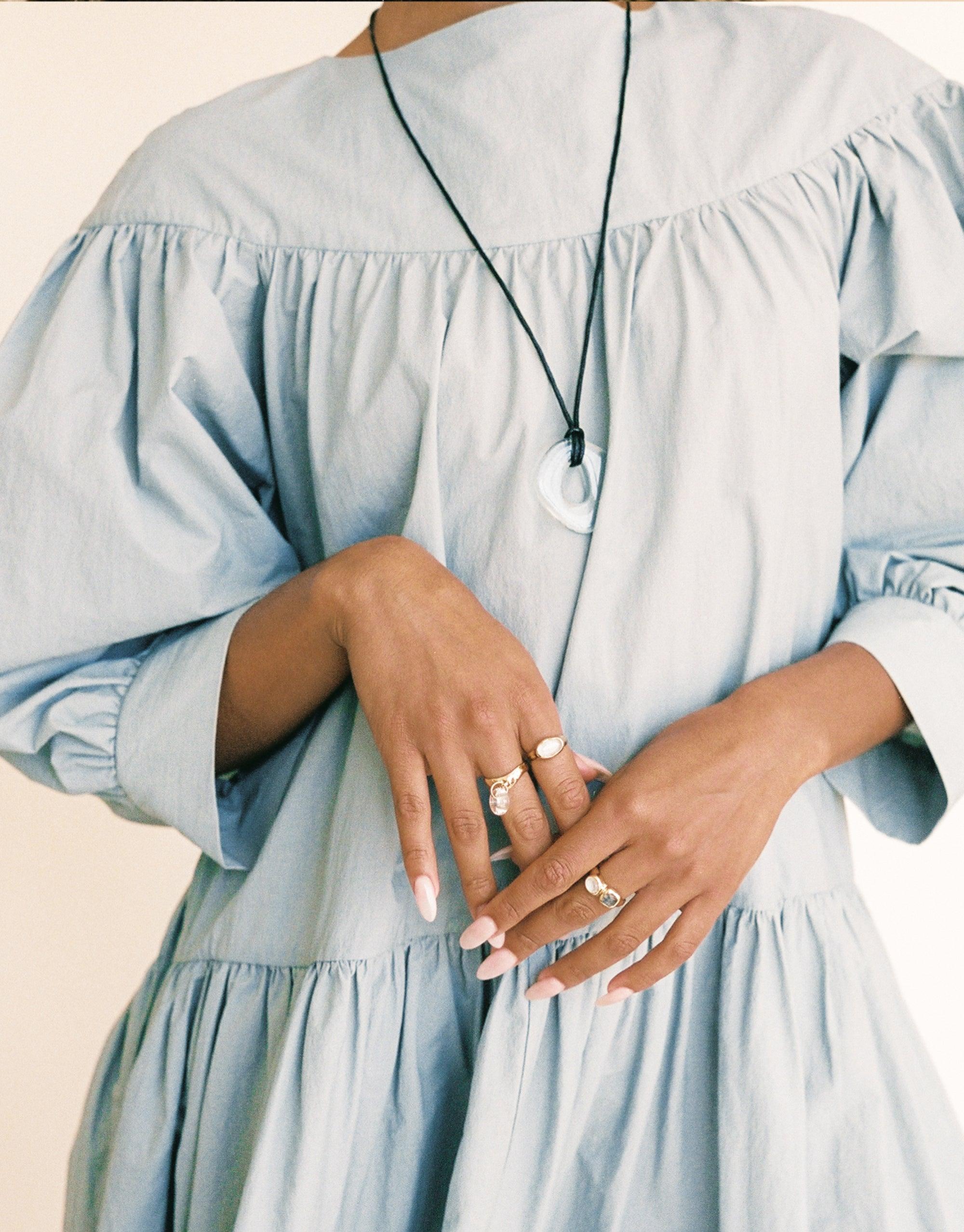 Close-up of hands wearing multiple rings, including the Duo Sculpture Ring, against a light blue ruffled dress. A white pendant necklace is visible. Showcases elegant jewelry styling and feminine fashion. CLED Duo Sculpture Ring.