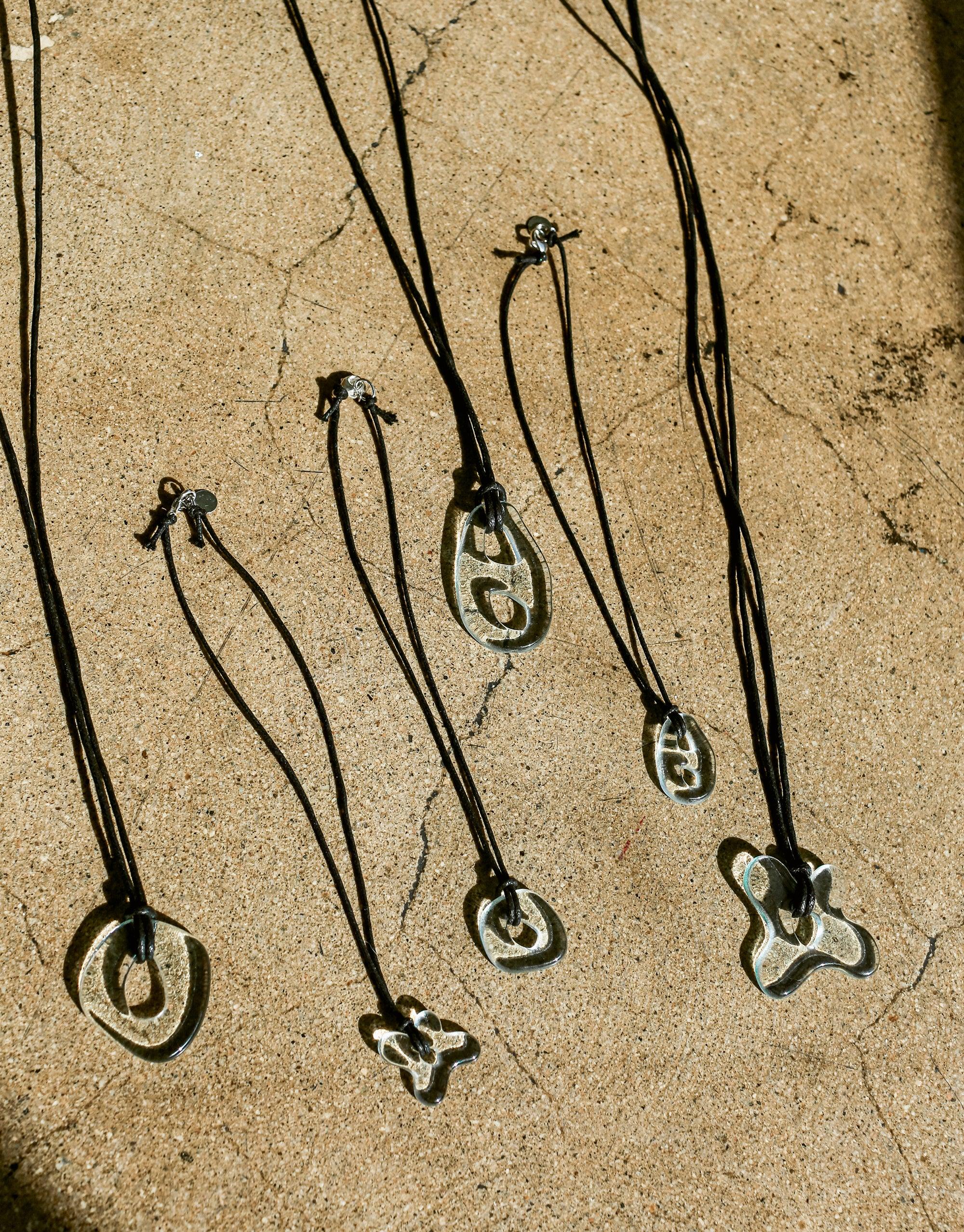 Sculptural Small Charm Necklace featuring unique 3D glass charms on black cotton cords. Various abstract shapes displayed on textured beige surface. Handcrafted eco-friendly jewelry with upcycled window glass. Sustainable and artistic design by CLED. Sculptural Small Charm Necklace | C