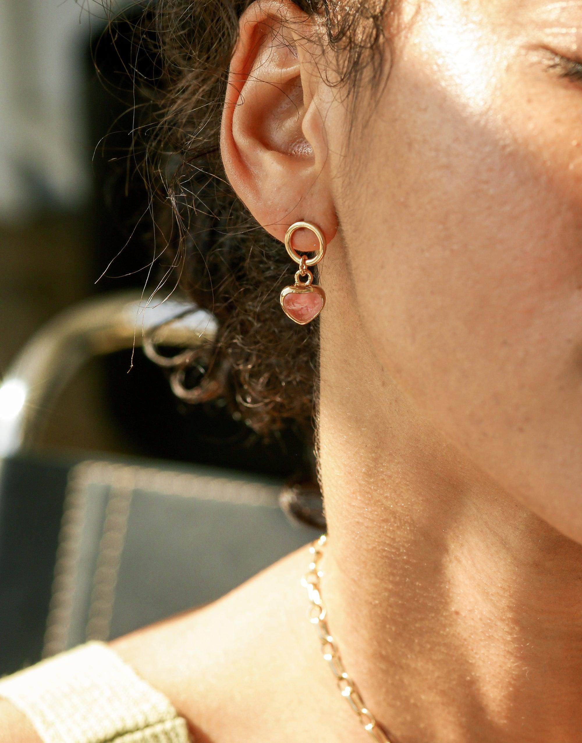 Close-up of a person wearing Heart Dangling Earrings, showcasing delicate gold hoop with a small pink heart charm. Sunlight highlights the earring's elegant design and the wearer's skin. Handcrafted jewelry by CLED.