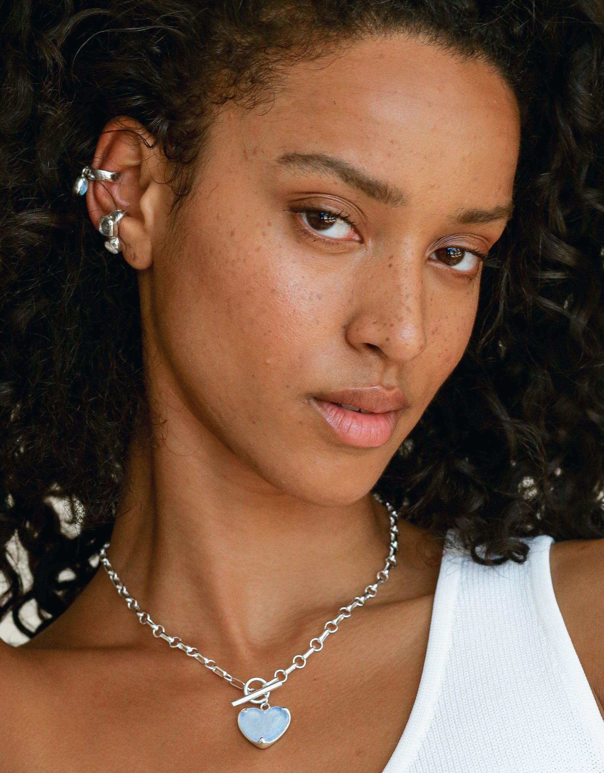 Close-up of ear wearing stylish Duo Sculpture Ear Cuff, featuring multiple charms and upcycled eco gems. Model also displays matching necklace with heart pendant. Handcrafted jewelry showcasing sustainable design and charm-inspired aesthetics. CLED Duo Sculpture Ear Cuff.