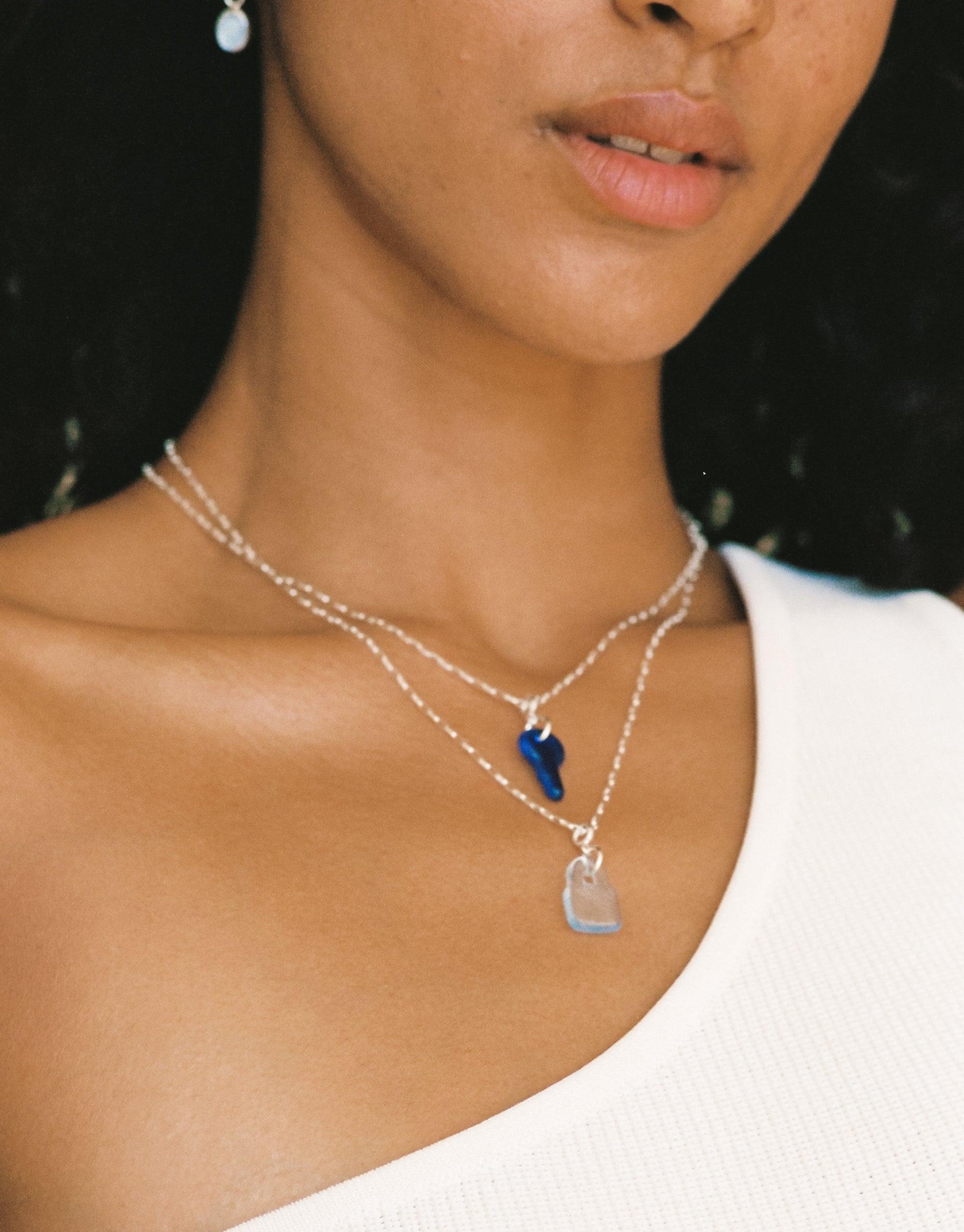 Close-up of model wearing layered silver necklaces featuring blue and clear Lock Icon Charms. Delicate chains showcase handcrafted eco-friendly pendants, highlighting the versatile and sustainable design of CLED's Lock | Icon Charm collection.