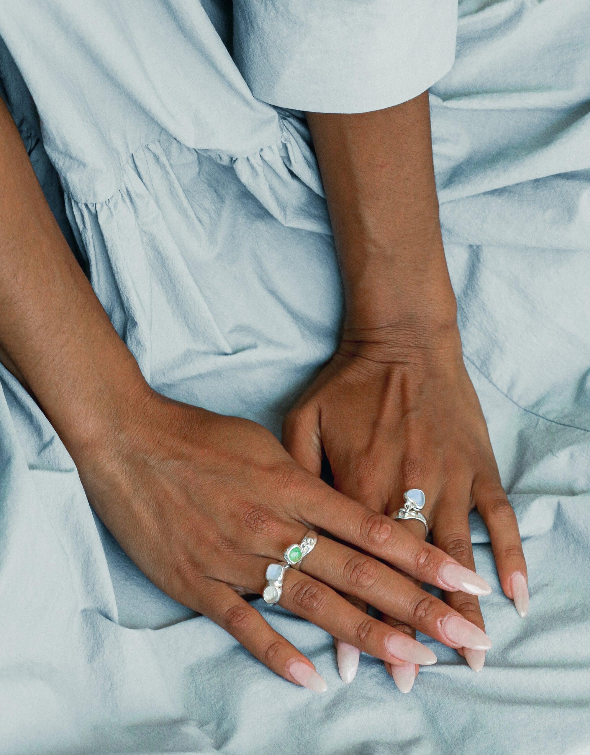 Close-up of hands with manicured nails wearing delicate silver rings, including a Heart Dangling Ring. The hands rest on light blue fabric, showcasing the elegant jewelry designs. Handcrafted, eco-friendly accessories by CLED.