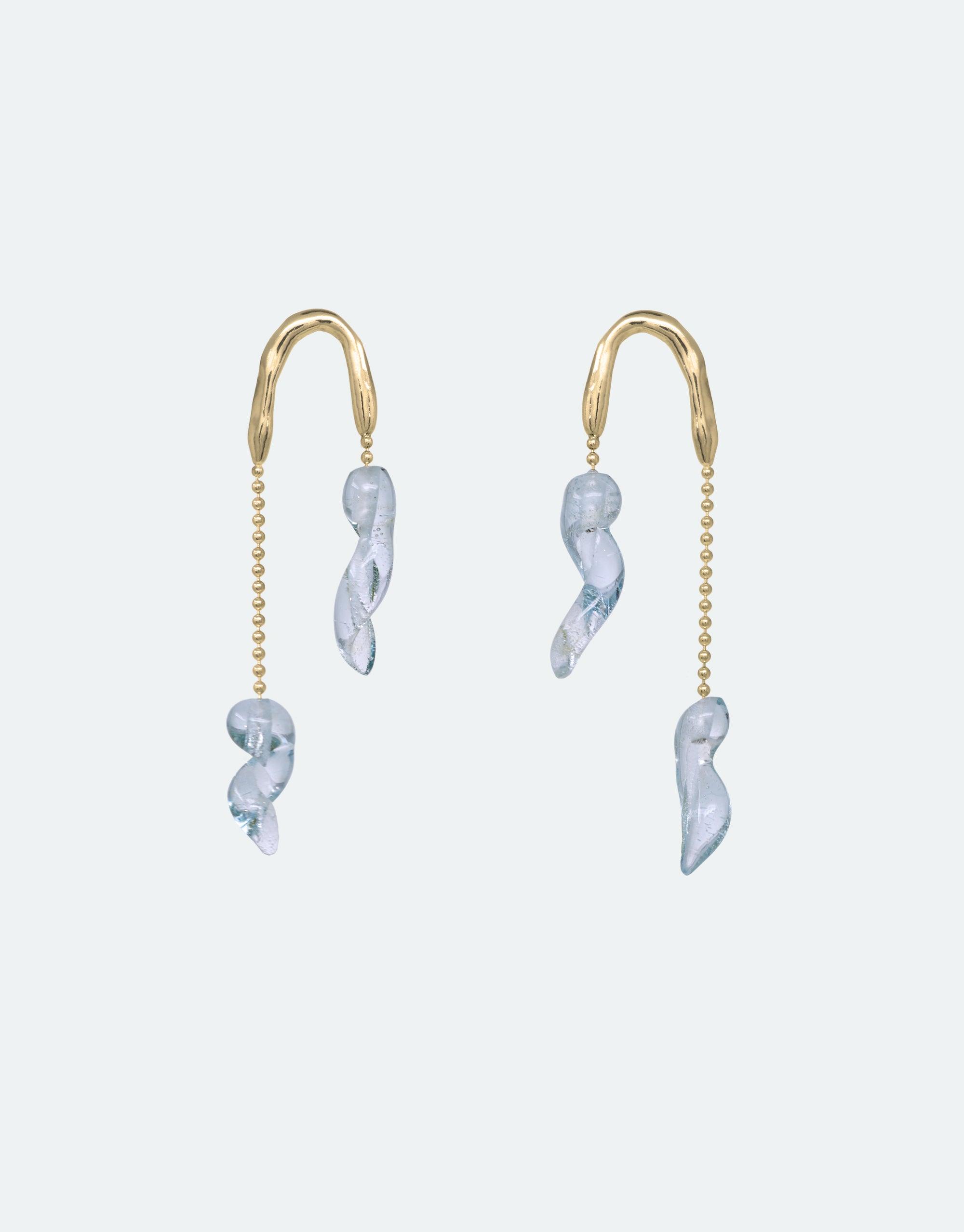 Asymmetrical gold earrings with delicate light blue glass drops resembling icicles, suspended on curved hooks and thin chains. Elegant design inspired by floating glaciers, showcasing eco-friendly materials. Crystal Drops Earrings by CLED.