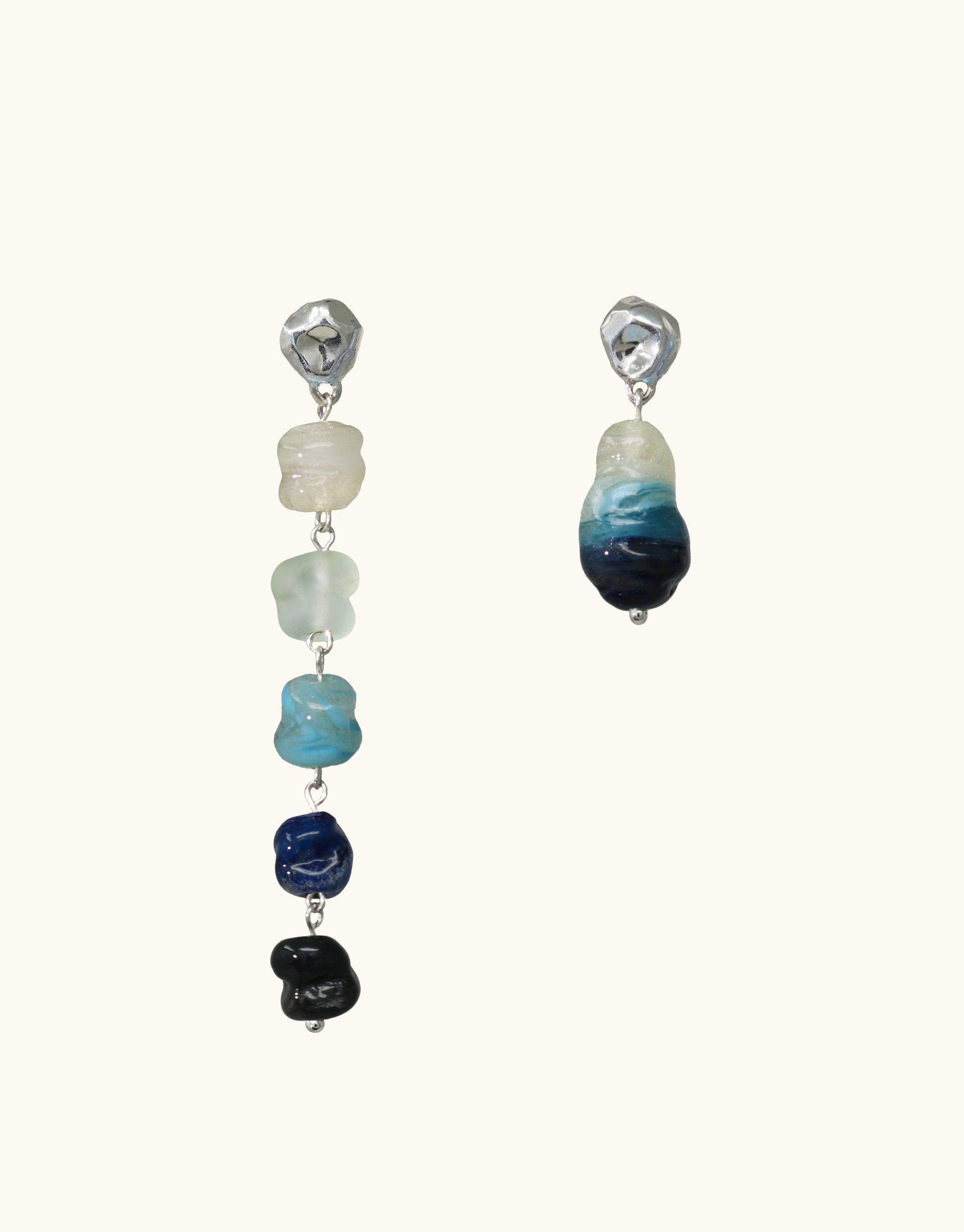 Asymmetrical earrings featuring eco-friendly glass beads in gradient blue shades, reminiscent of cascading waterfalls. Long earring with multiple beads, short earring with single bead. Handcrafted sustainable jewelry. Shades of Nature | Waterfall Earrings by CLED.