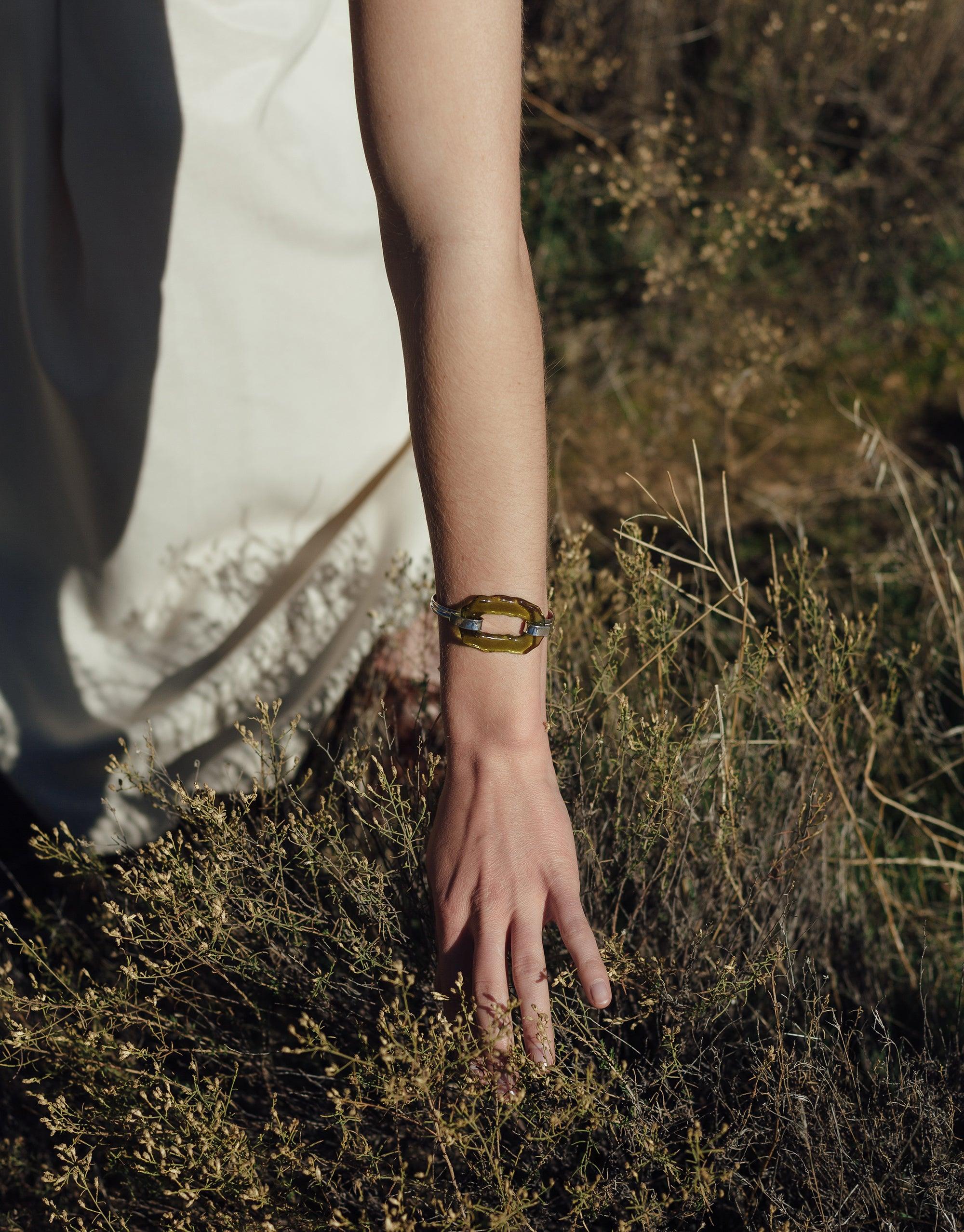 Close-up of a hand wearing the Canyon Hook Bracelet against a natural, outdoor backdrop with dry grass and foliage, showcasing the bracelet's organic oval shape and eco-friendly design inspired by canyon terrain. CLED Canyon Hook Bracelet.