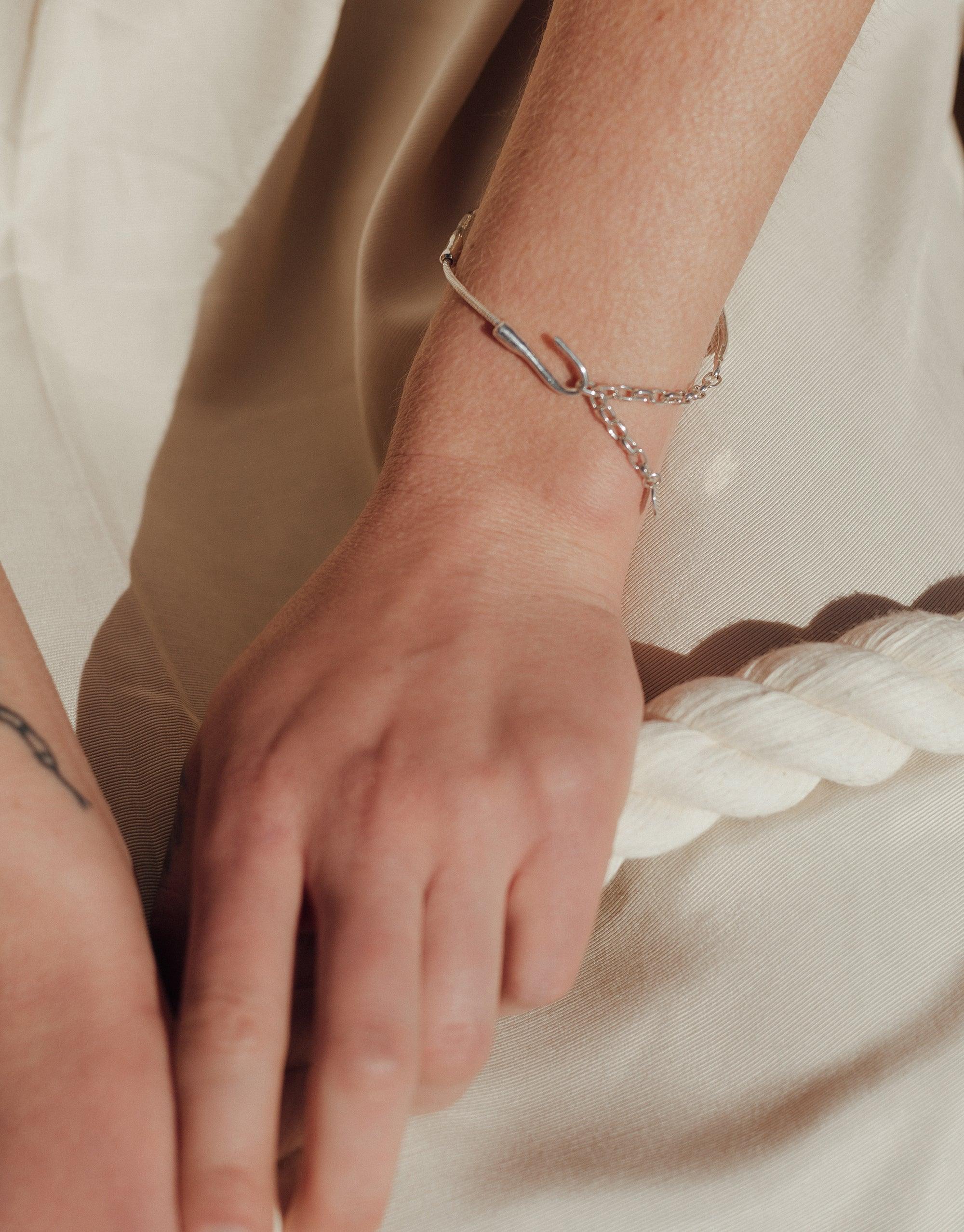 Close-up of a wrist wearing a sleek silver Single Rope Hook Bracelet. The bracelet features a minimalist design with a hook closure and delicate chain, showcasing the edgy yet elegant style of the Terrain Collection. The bracelet rests against light-colored fabric, emphasizing its refined craftsmanship. CLED Single Rope Hook Bracelet.