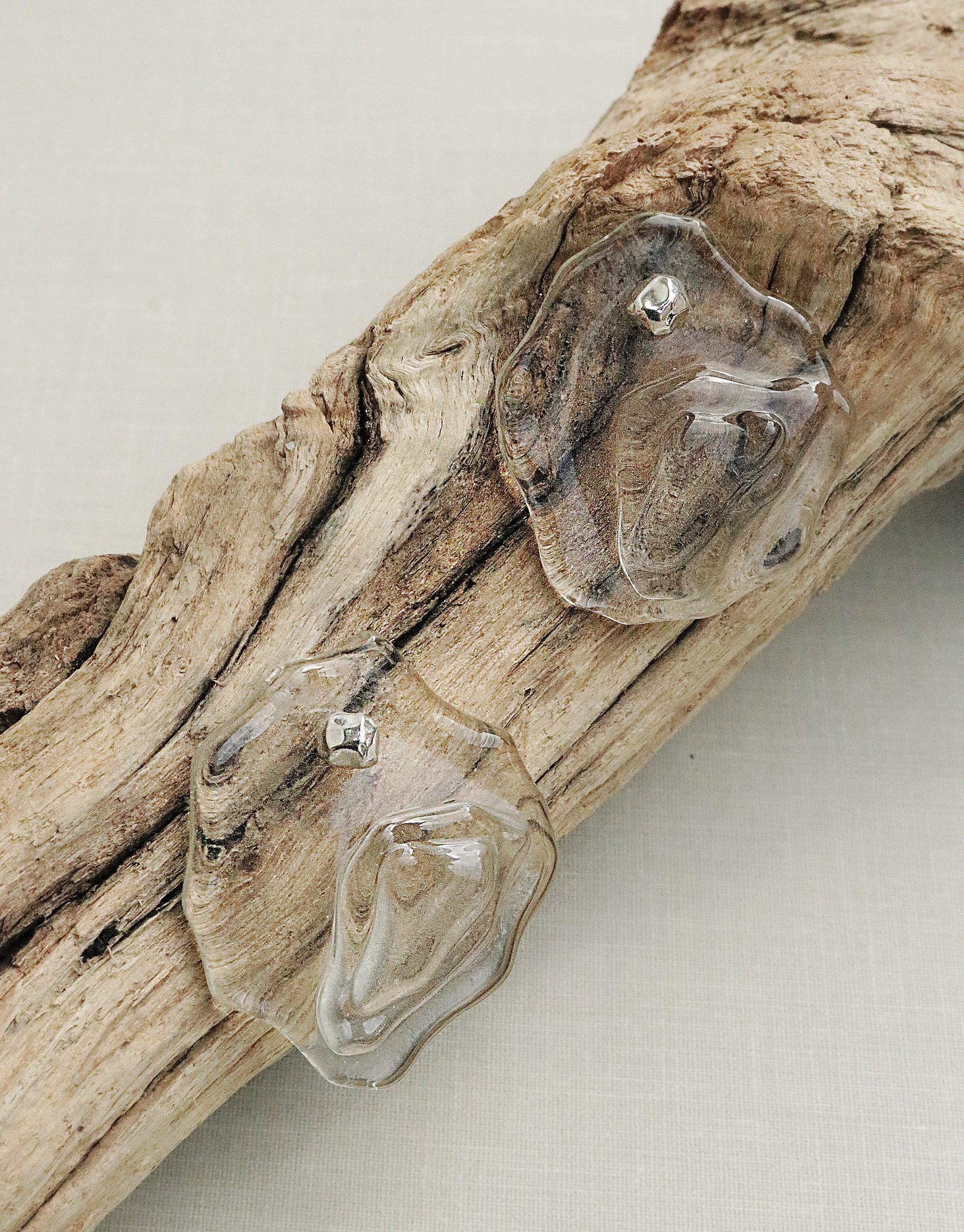 Plateau Earrings displayed on driftwood, featuring abstract molten glass forms inspired by canyon terrain. Handcrafted eco-friendly jewelry with layered transparent elements and silver accents. Sustainable, unique design showcasing natural textures and shapes. CLED Plateau Earrings.