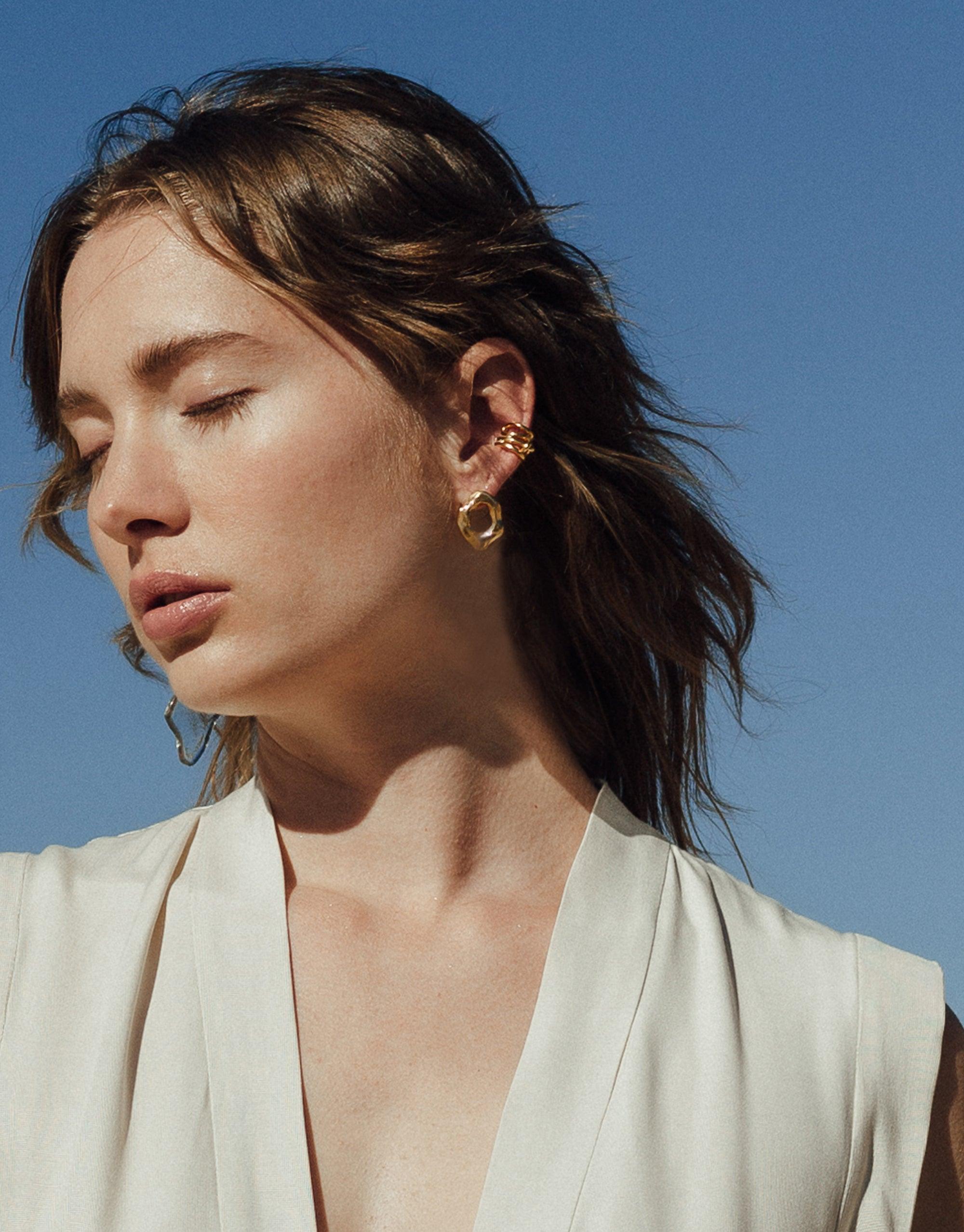 Model wearing Canyon Stud Earrings, showcasing abstract molten forms inspired by canyon terrain. Gold vermeil earrings displayed on ear against bright blue sky background. Handcrafted jewelry from Terrain Collection by CLED.