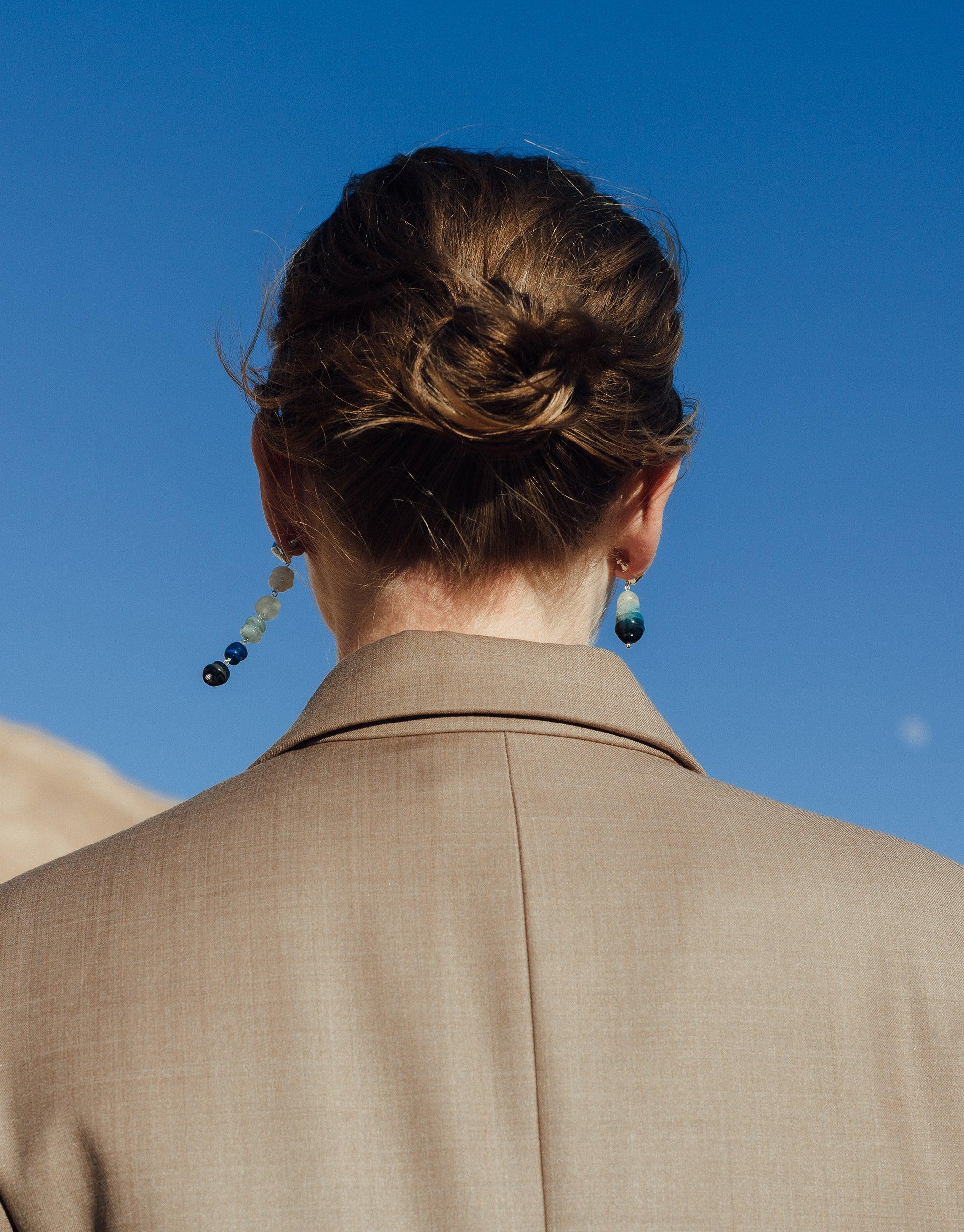 Back view of person wearing Shades of Nature Waterfall Earrings, showcasing long and short blue eco-glass designs against beige coat and clear sky, highlighting canyon-inspired Terrain Collection. Handcrafted sustainable jewelry by CLED.