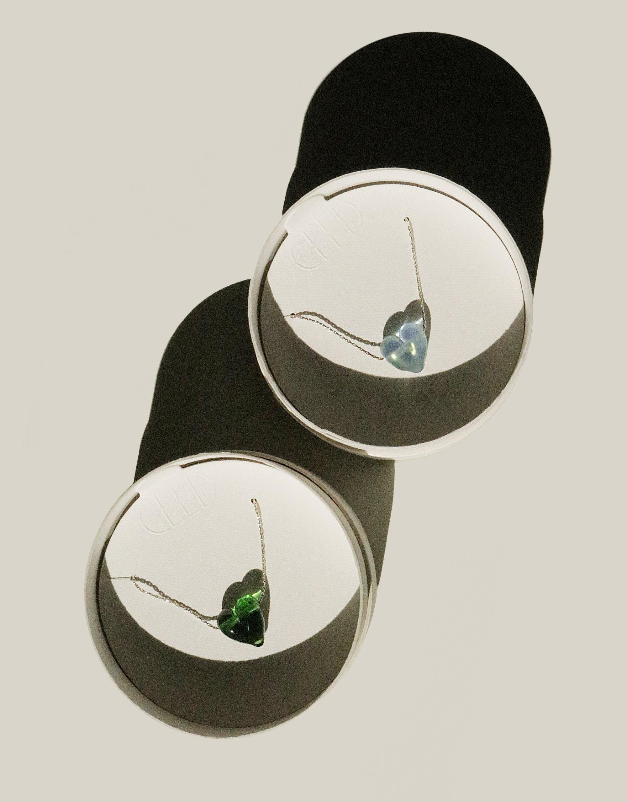 Two circular frames showcasing Love Necklaces with heart-shaped pendants in blue and green, made from repurposed glass on delicate chains. Elegant and sustainable jewelry design by CLED.