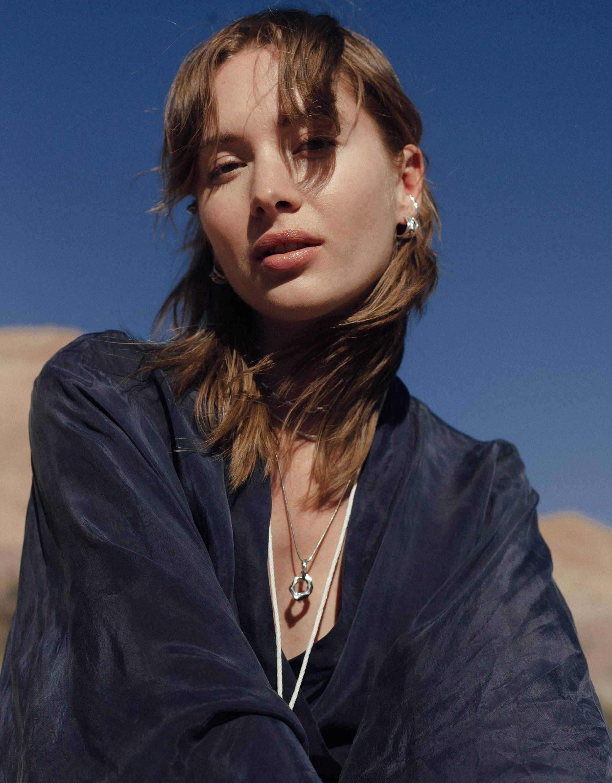 Model wearing Canyon Necklace from Terrain Collection, featuring abstract molten pendant against desert backdrop. Sleek design showcases smooth curves in sterling silver or gold vermeil, perfect for layering. Handcrafted 20-inch necklace inspired by canyon formations. CLED Canyon Necklace.