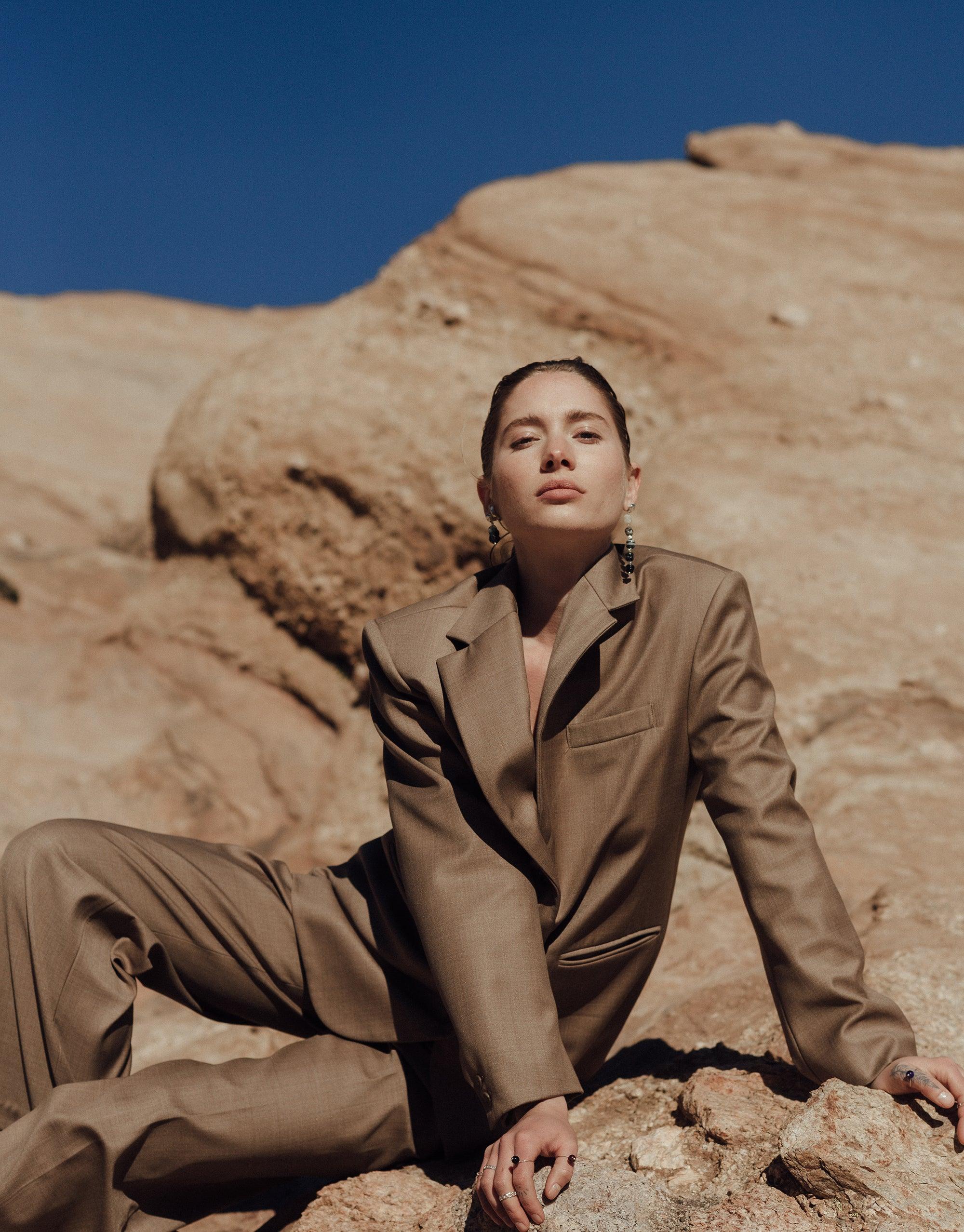 Model wearing Shades of Nature Basalt Earrings in desert canyon setting, showcasing eco-friendly jewelry inspired by volcanic terrain. Elegant long earrings complement tan suit, highlighting sustainable fashion against rocky backdrop. CLED Shades of Nature | Basalt Earrings