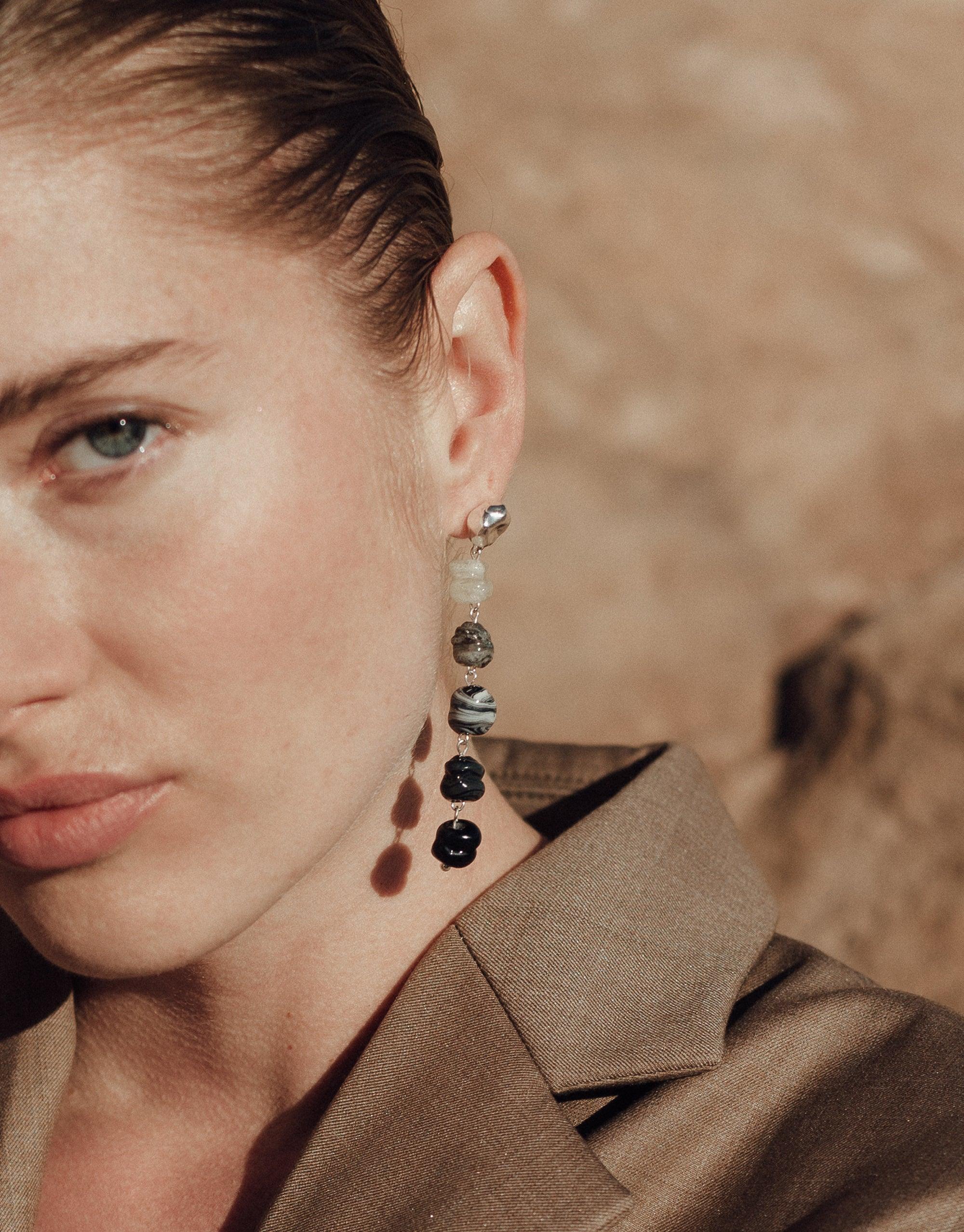 Close-up of model wearing Shades of Nature Basalt Earrings, showcasing unique black and white glass beads in varying sizes, inspired by volcanic terrain. Earrings hang elegantly from ear, complementing natural-toned background. Eco-friendly jewelry by CLED.