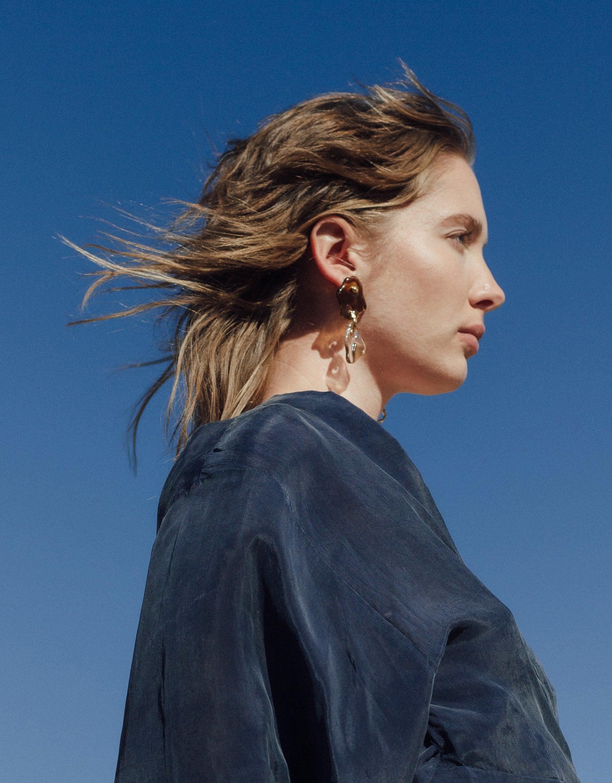 Profile view of model wearing Peak Earrings against clear blue sky. Long, windswept hair. Earrings feature layered glass charms inspired by canyon terrain. Handcrafted sustainable jewelry from Terrain Collection. Peak Earrings by CLED.