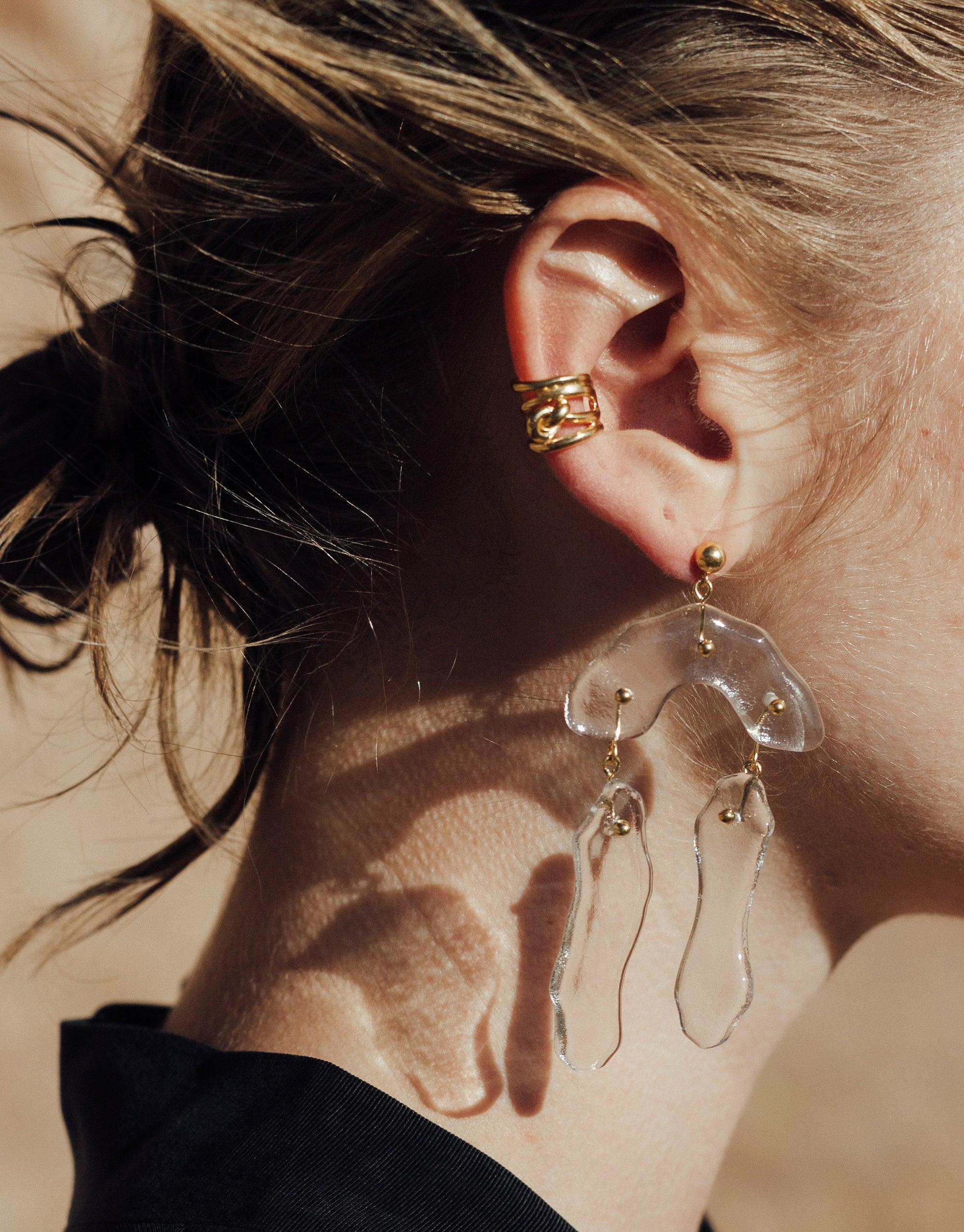Close-up of a model wearing a gold Rope Ear Cuff on the upper ear, paired with a statement glass earring featuring abstract shapes. The image showcases the versatility of the Rope Ear Cuff | Single, highlighting its ability to complement other jewelry pieces from the Terrain Collection by CLED.