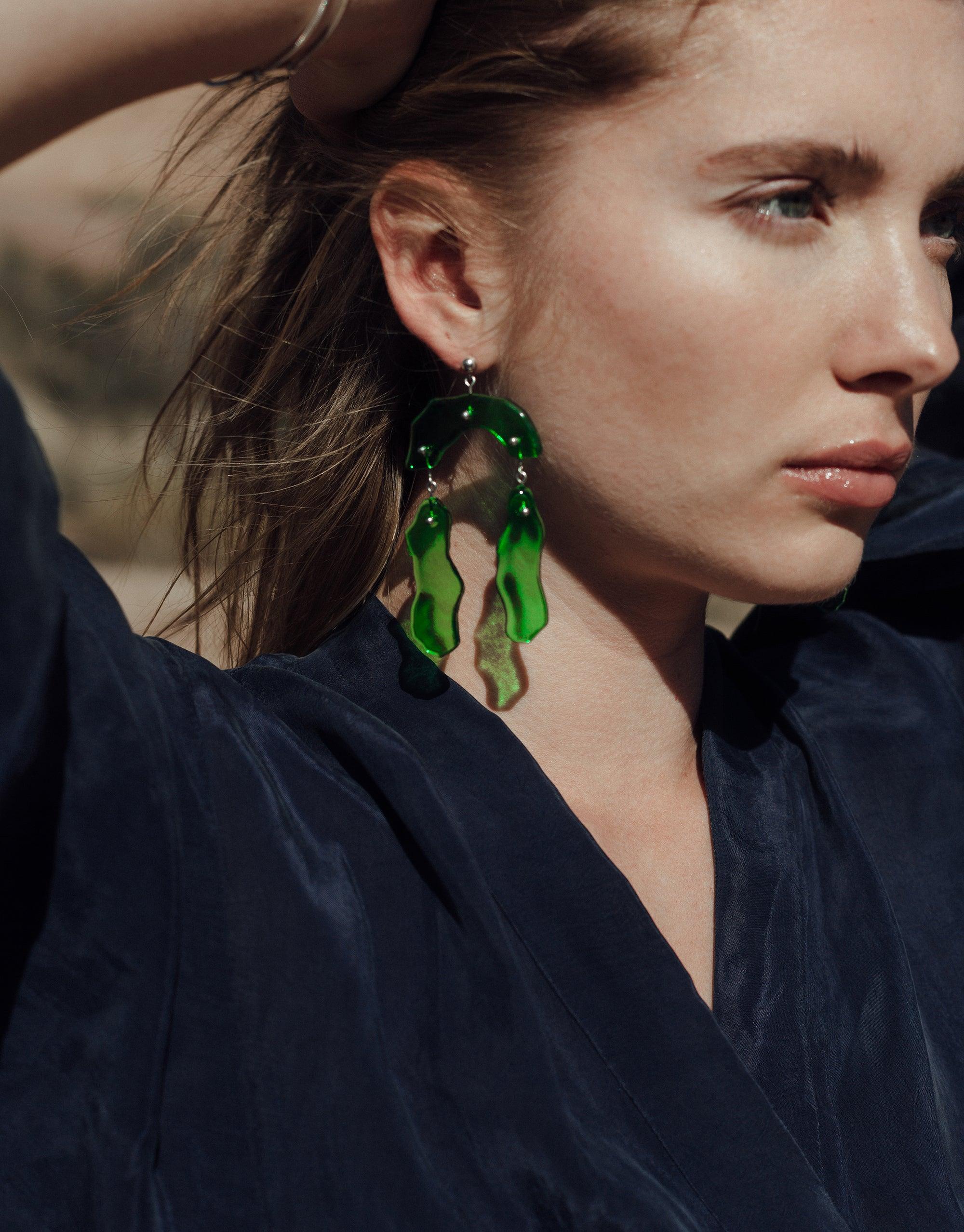 Close-up of person wearing unique green glass dangling earrings with abstract shapes, reminiscent of canyon terrain. Showcases Rock Dangling Earrings from the Terrain Collection, highlighting eco-friendly design and artisanal craftsmanship. CLED