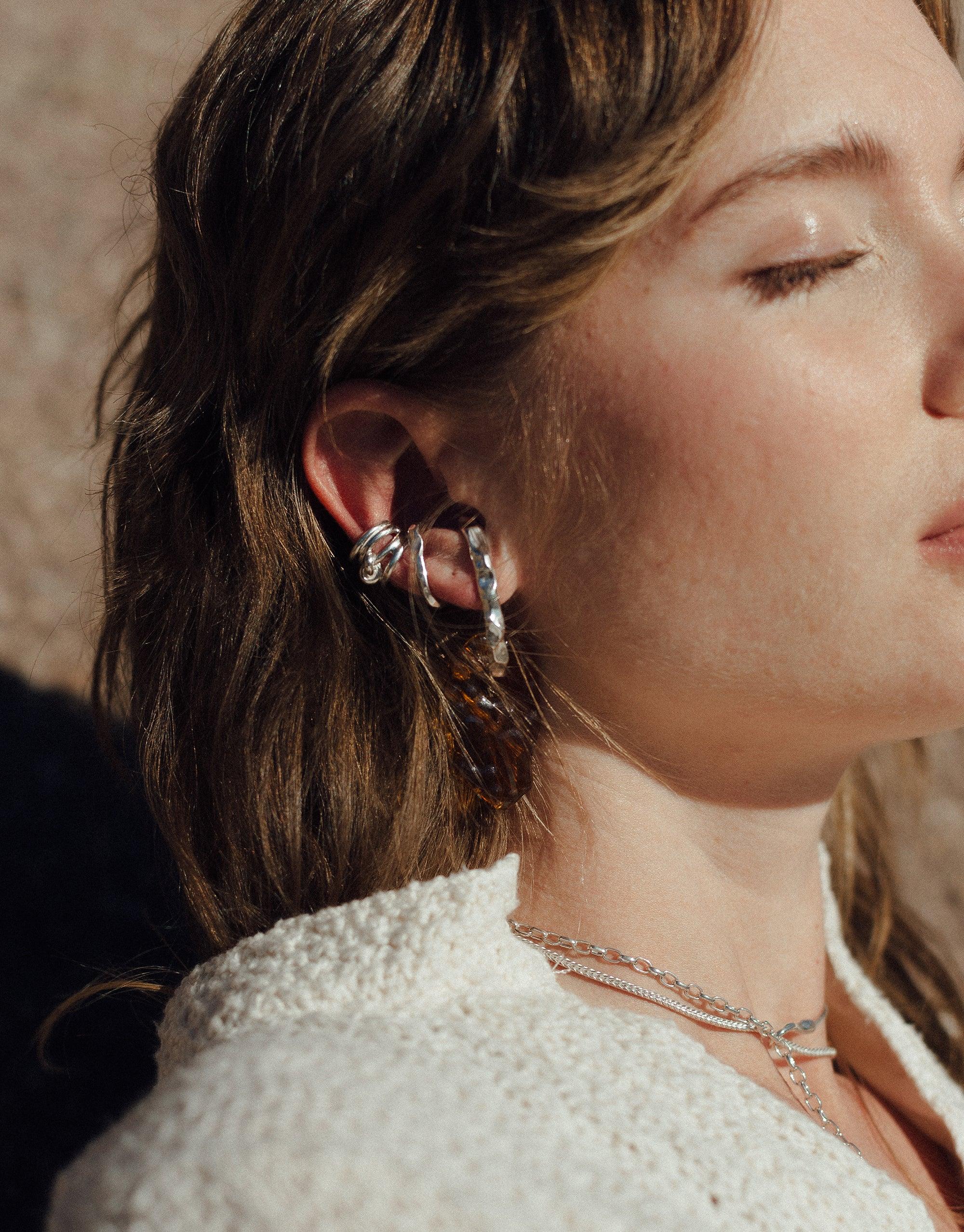 Close-up of a person wearing multiple ear cuffs and earrings, showcasing the Rock Ear Cuff | Single. The jewelry pieces have a unique, abstract design inspired by canyon terrain. The image highlights the versatility and stylish nature of CLED's Terrain Collection.
