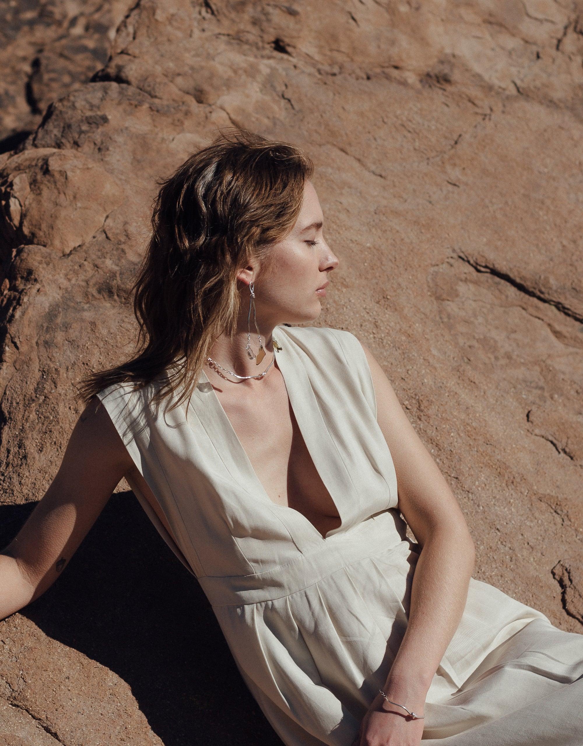 Woman in white dress sitting against rugged canyon rock formation, showcasing Rope Drop Earrings inspired by terrain and climbing equipment. Eco-friendly jewelry by CLED.