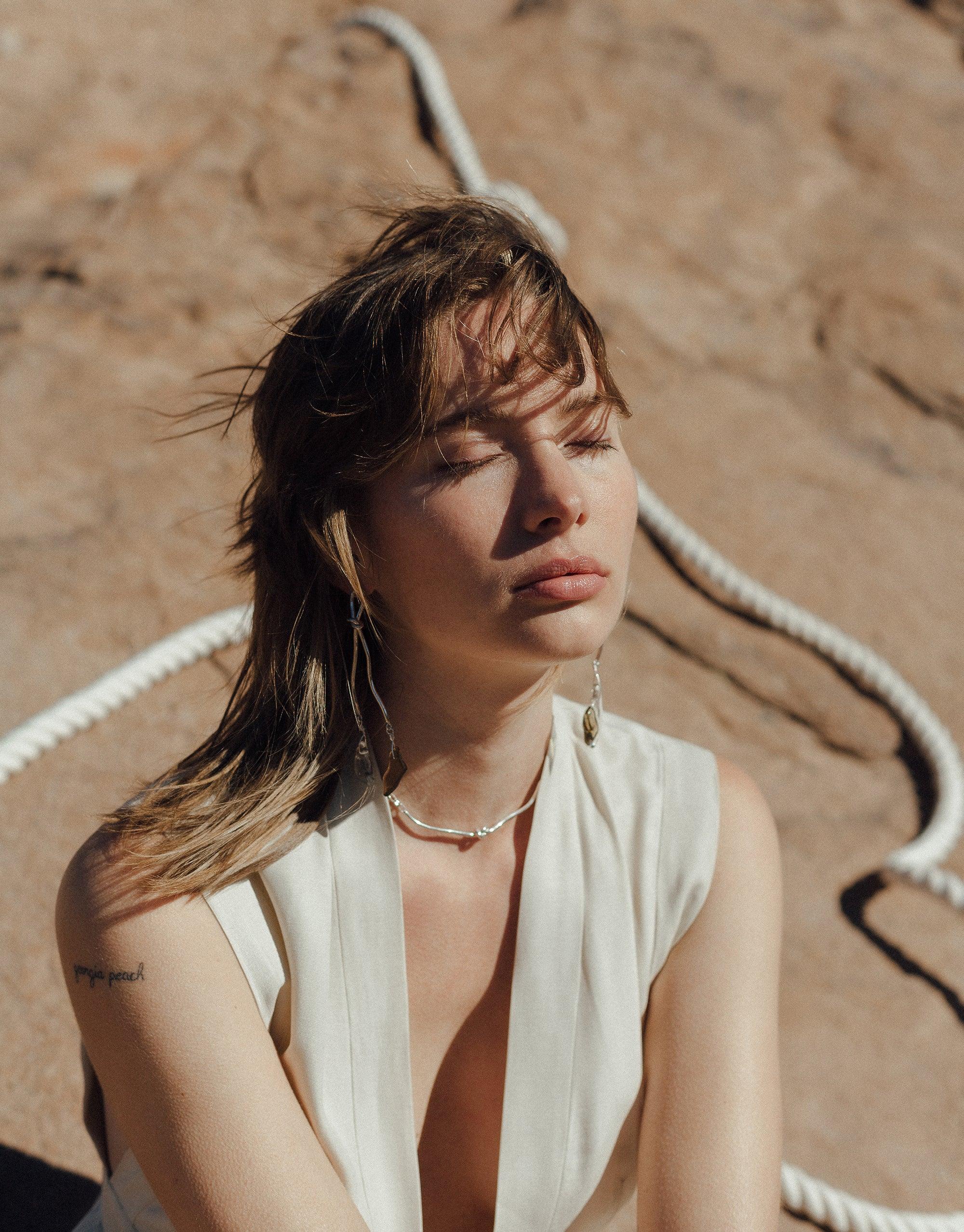 Woman in white top wearing elegant canyon-inspired Rope Drop Earrings against desert backdrop, showcasing handcrafted eco-friendly jewelry from Terrain Collection. Sunlight highlights unique design and sustainable materials. Rope Drop Earrings by CLED.