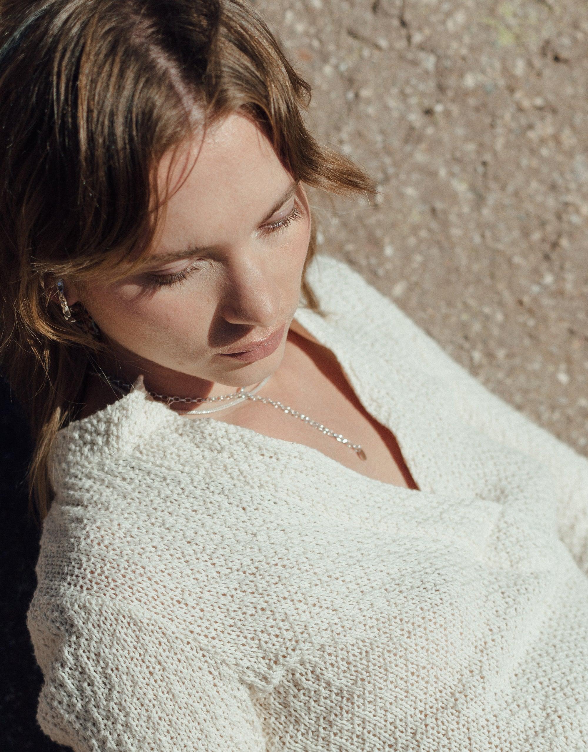 Close-up of person wearing white knit sweater and delicate Rope Hook Necklace, showcasing the necklace's elegant design against sun-kissed skin. Inspired by canyon terrain, this versatile piece from the Terrain Collection by CLED can be worn in front or back.