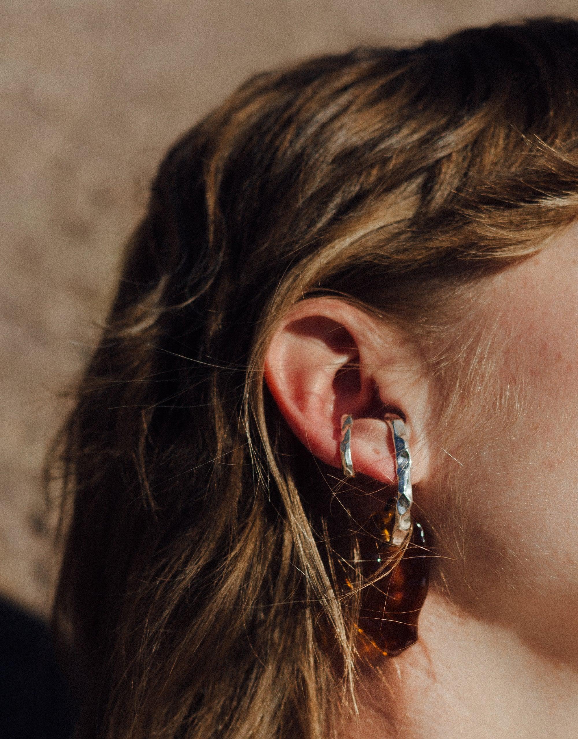 Close-up of model wearing Rock Spire Earrings, showcasing curved metallic design with glass drop, part of Terrain Collection inspired by canyon formations. Handmade eco-friendly statement piece by CLED.