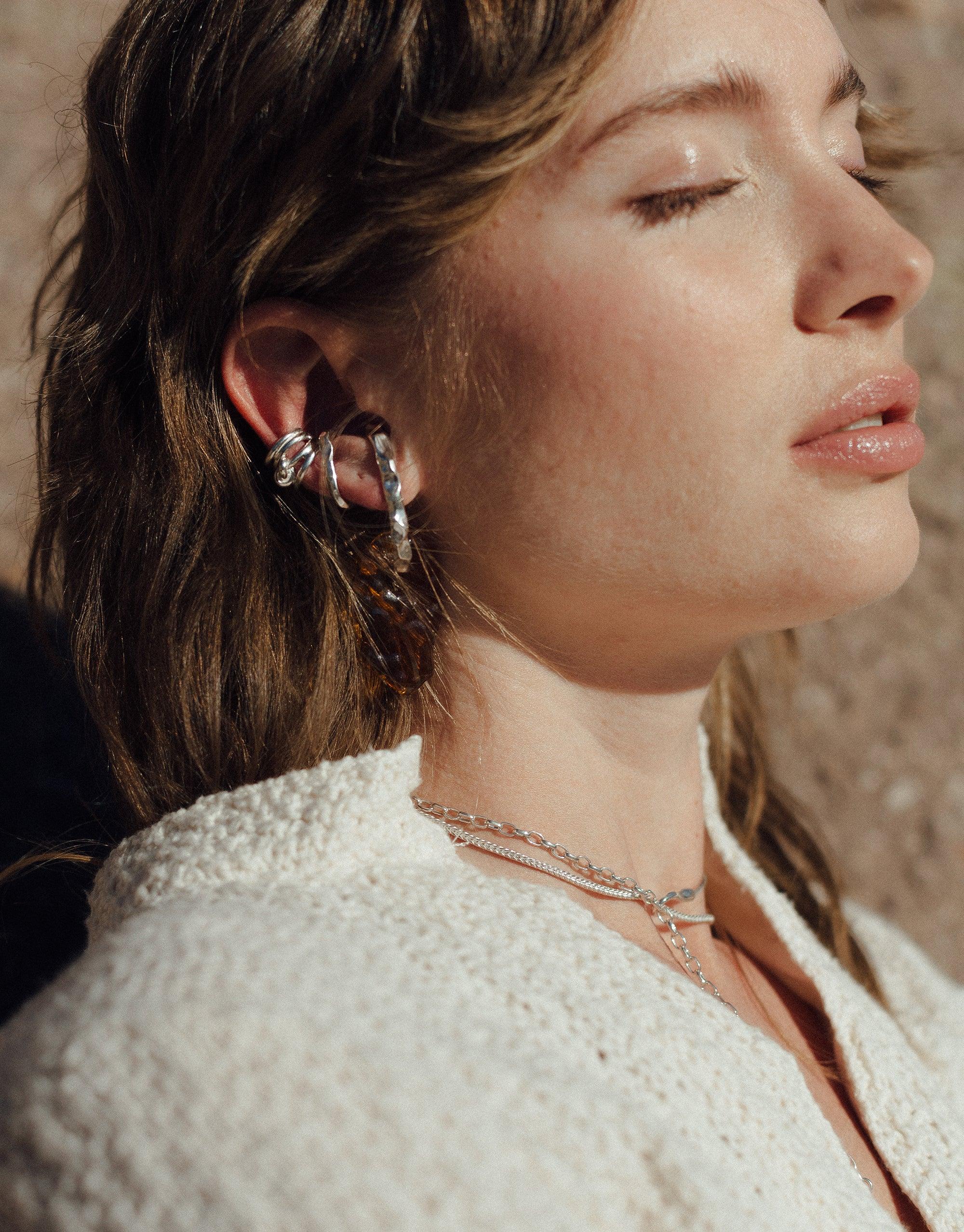 Close-up profile of a woman wearing a delicate Rope Hook Necklace, showcasing its elegant design against her neck. The necklace complements her textured white top, embodying the abstract molten forms inspired by canyon terrain. Terrain Collection by CLED.