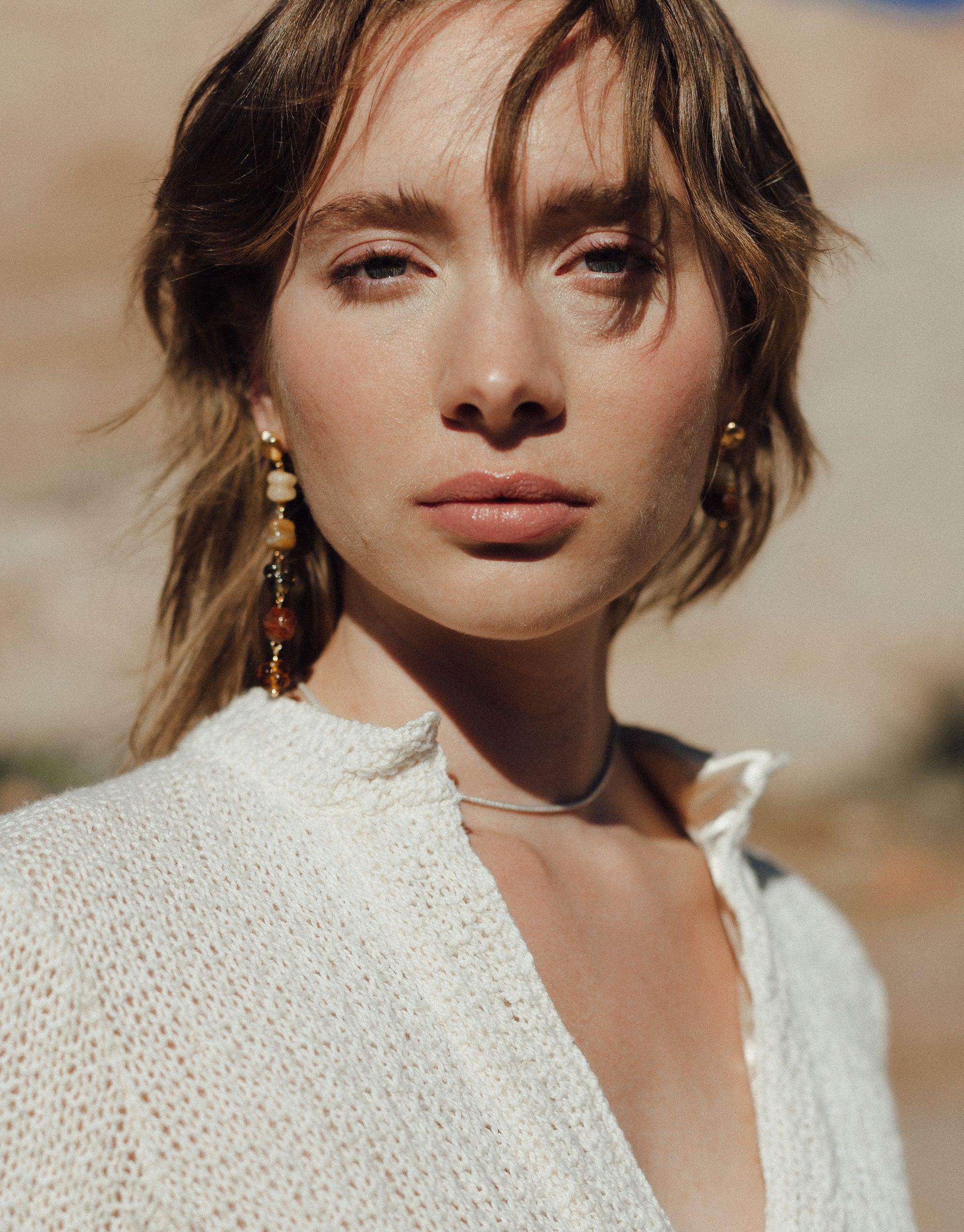 Elegant model wearing Shades of Nature Sandstone Earrings, showcasing long dangling design inspired by canyon terrain. Eco-friendly glass earrings in warm earth tones complement white knit top. Handcrafted sustainable jewelry by CLED.