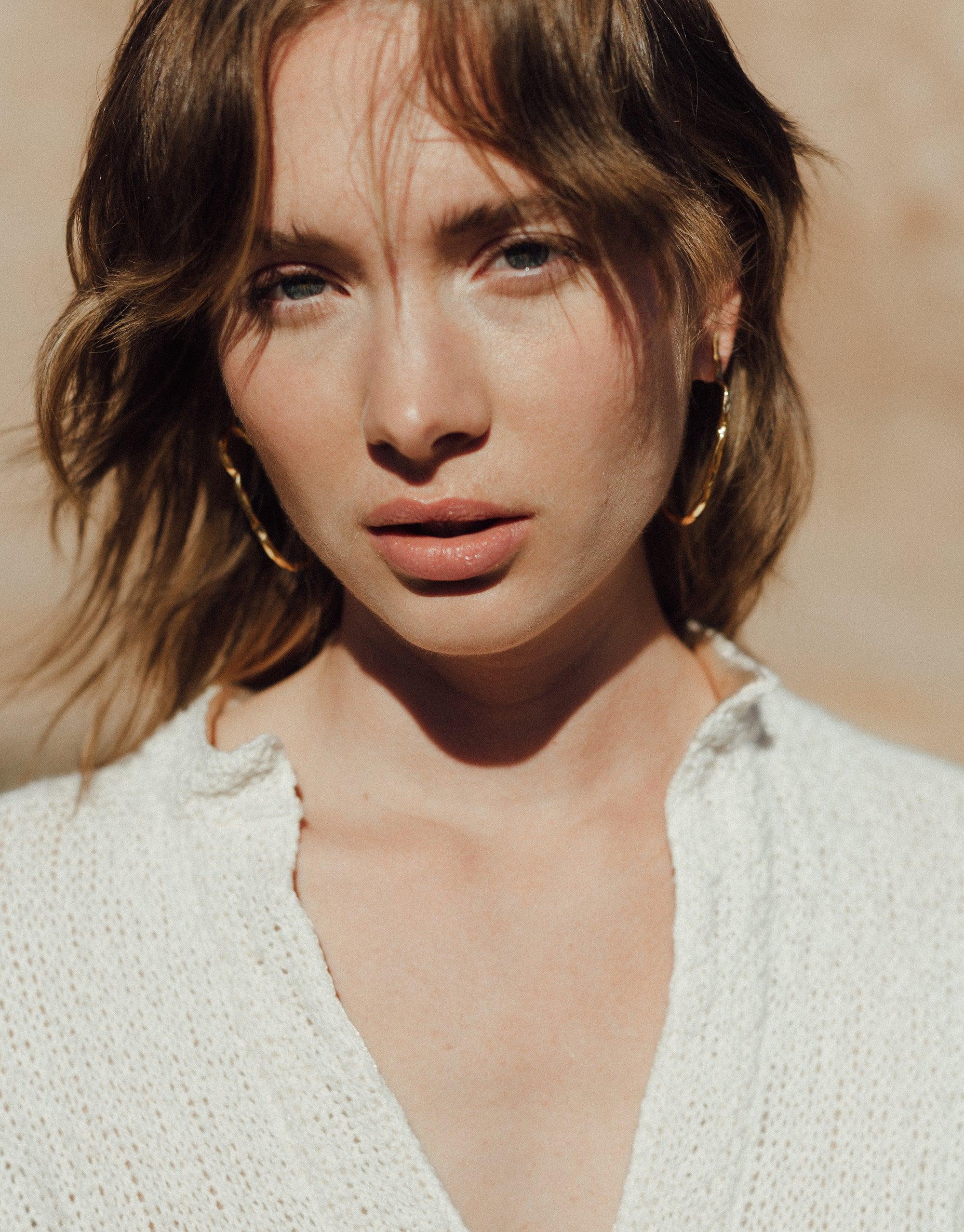 Model wearing Rock Stud Earrings, showcasing curved gold hoops against sunlit skin and tousled hair. Earrings complement white textured top, capturing essence of Terrain Collection inspired by canyon formations. Handcrafted jewelry by CLED.