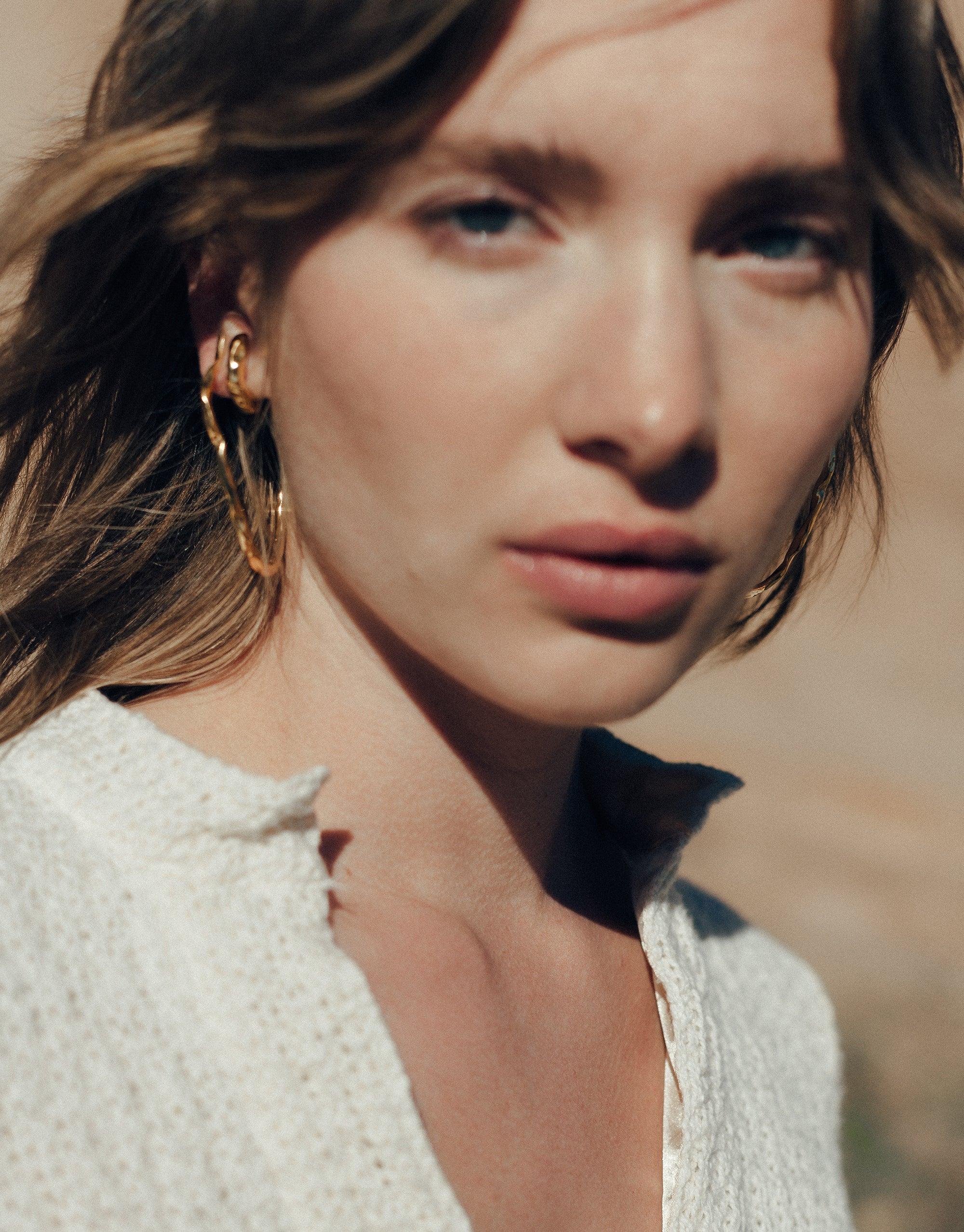Vale Hoop Earrings: A woman models open back hoop earrings with curved lines, inspired by canyon valleys, highlighting the Terrain Collection's abstract design, made in Los Angeles from eco-conscious materials.
