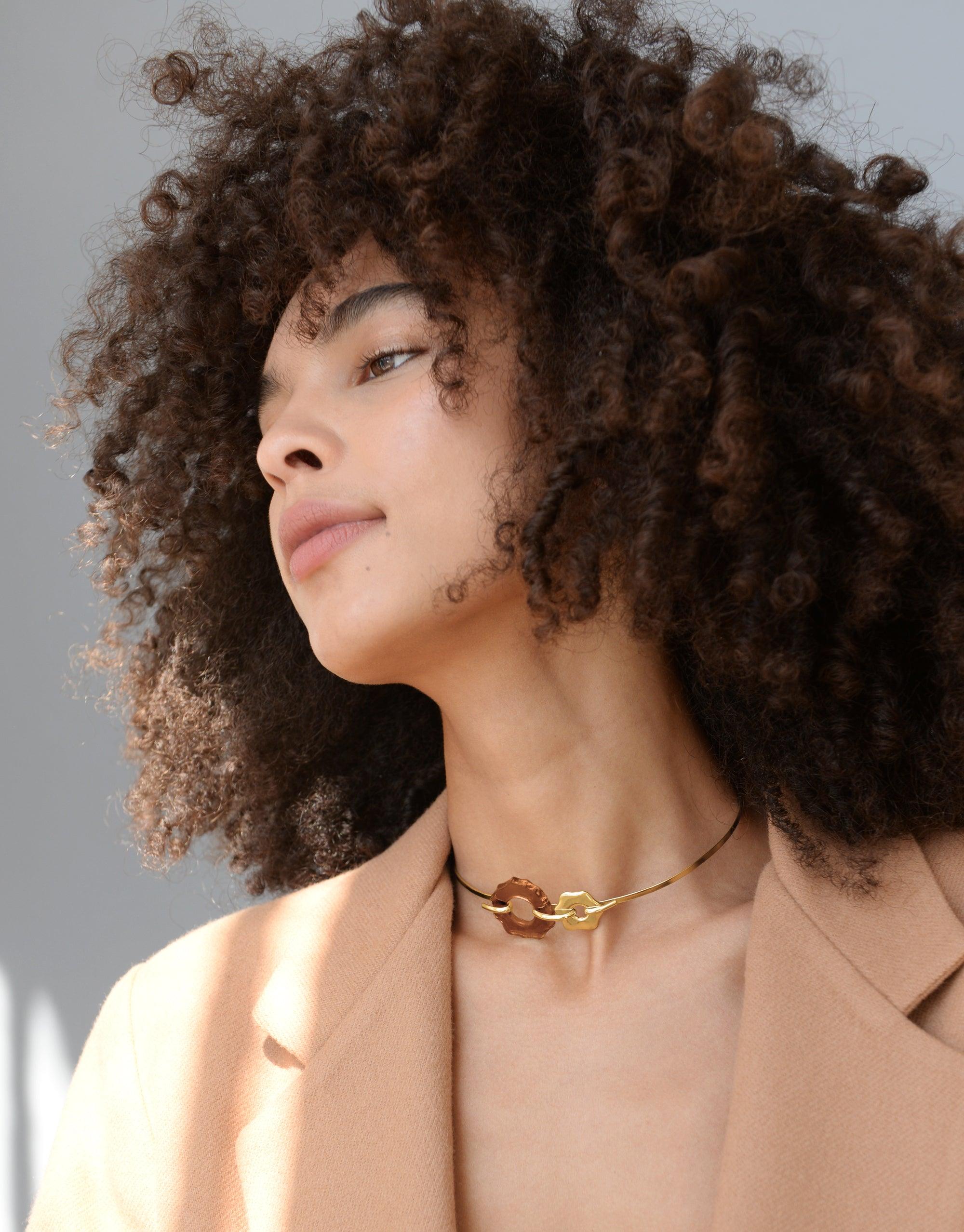 Profile view of model with voluminous curly hair wearing a peach blazer and the Avens Choker Necklace, featuring a delicate gold chain with petal-inspired glass droplets. Elegant and eco-conscious accessory showcasing CLED's commitment to sustainable fashion.