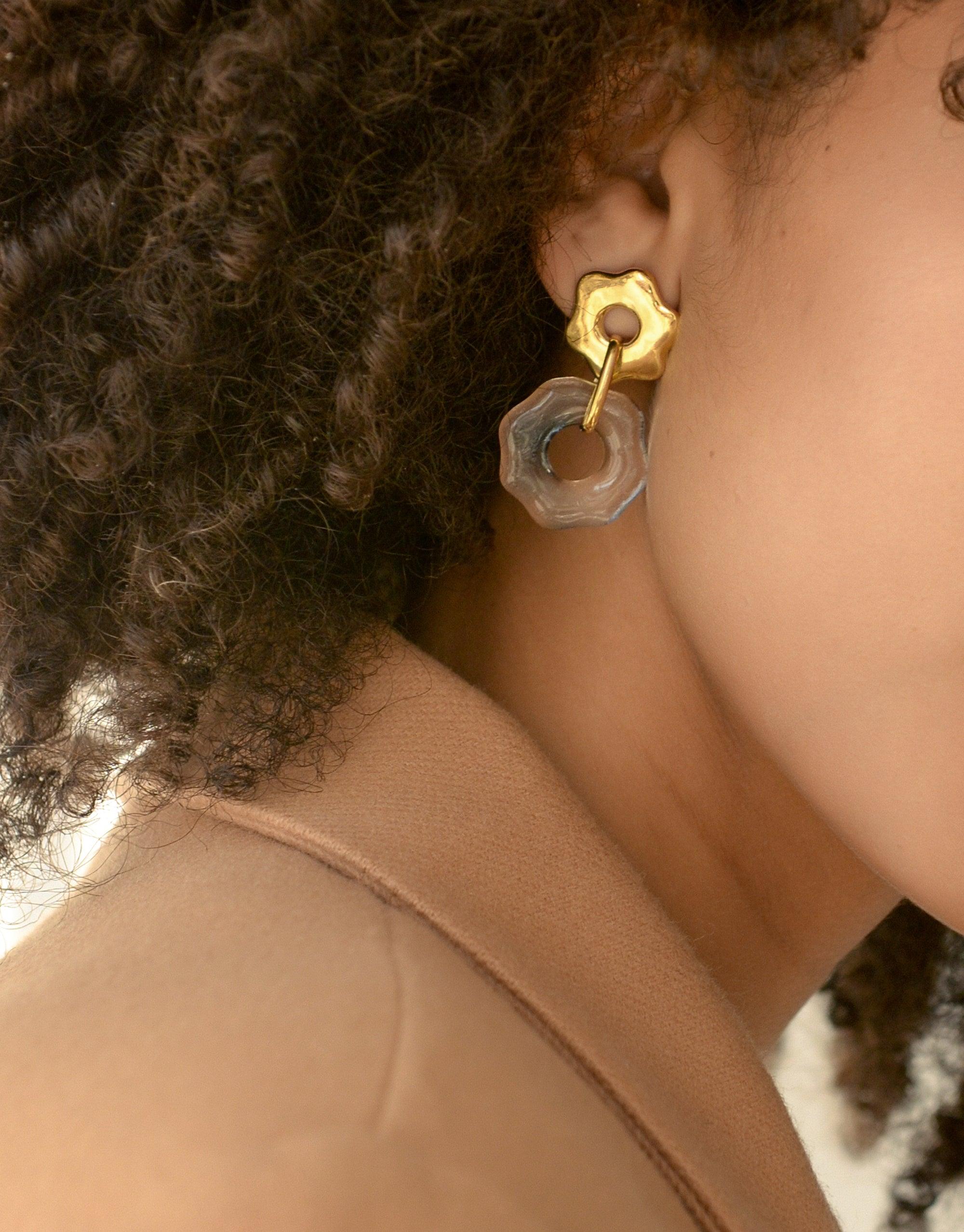 Avens Earrings: Gold flower-shaped stud with transparent octagonal pendant, showcasing eco-friendly design. Elegant and sustainable jewelry piece worn by model with curly hair. Inspired by Arctic flora and climate awareness. Handcrafted by CLED.