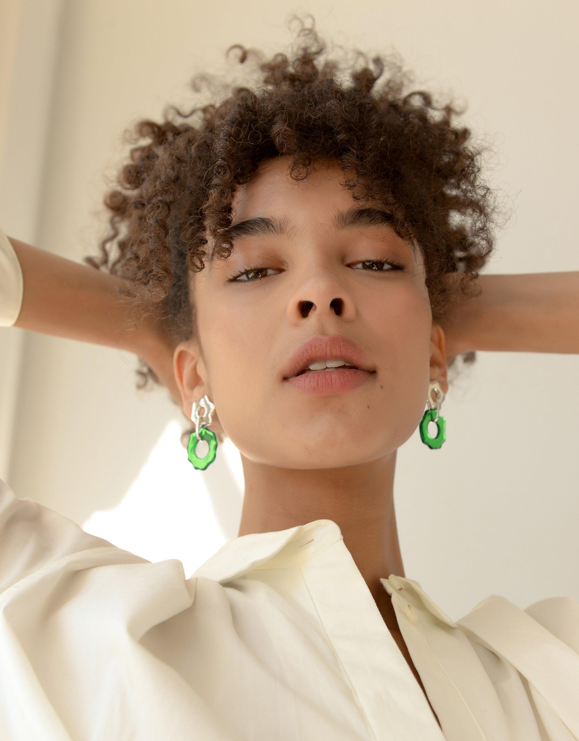 Close-up of a model wearing Avens Earrings, featuring vibrant green eco-gem drops made from repurposed glass. The earrings complement the model's natural curly hair and cream-colored top, showcasing the elegant design inspired by Arctic flora. Sustainable jewelry by CLED.