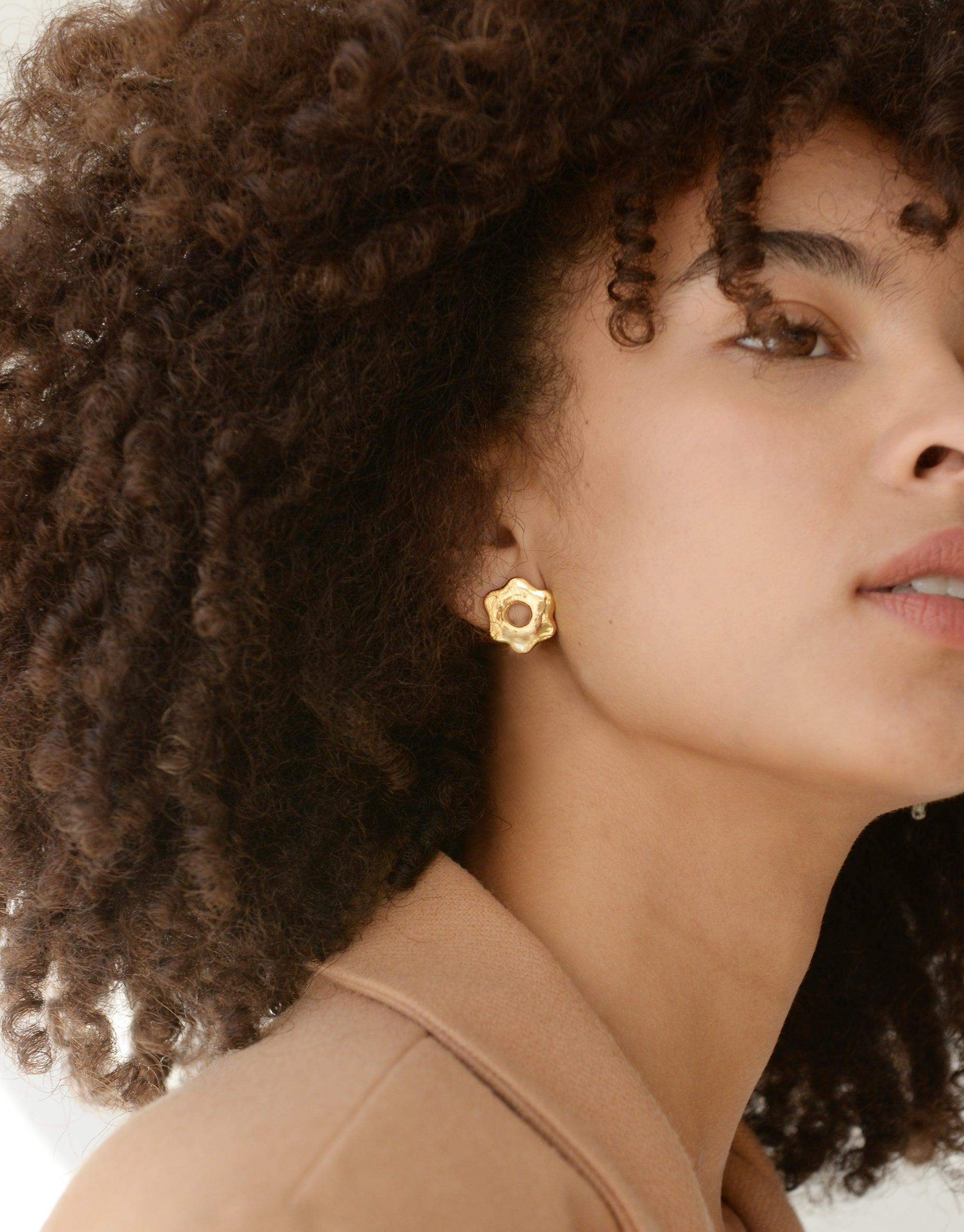 Close-up profile of a model with curly hair wearing the Avens Stud Earrings, showcasing the gold vermeil flower-shaped design against warm skin tone. Sustainable jewelry by CLED.