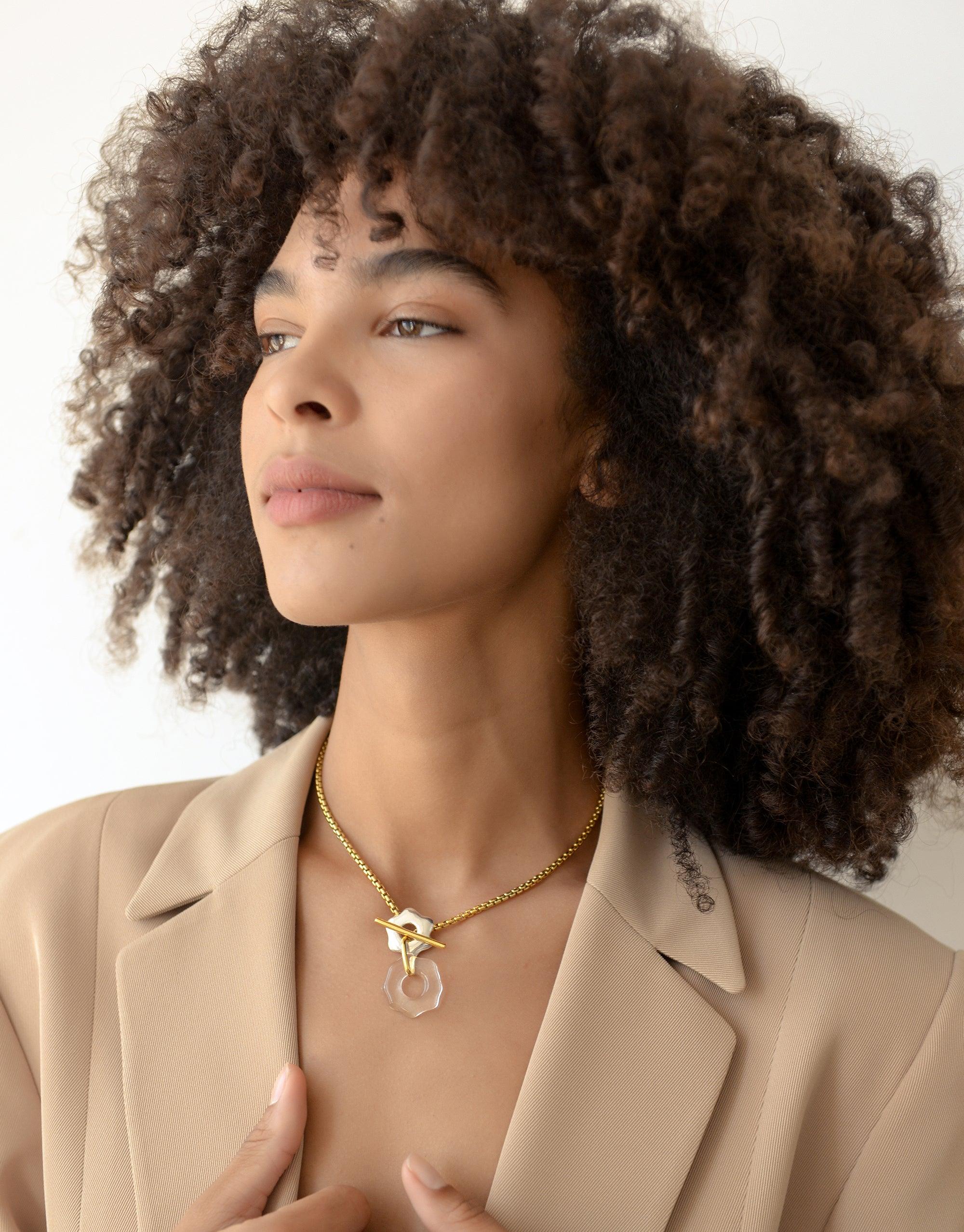 Close-up of a model wearing the Avens Toggle Necklace, featuring a gold chain with a clear pendant. The necklace sits elegantly against a beige blazer, showcasing the Floating Glacier Collection's eco-friendly design. Sustainable jewelry by CLED, Avens Toggle Necklace.