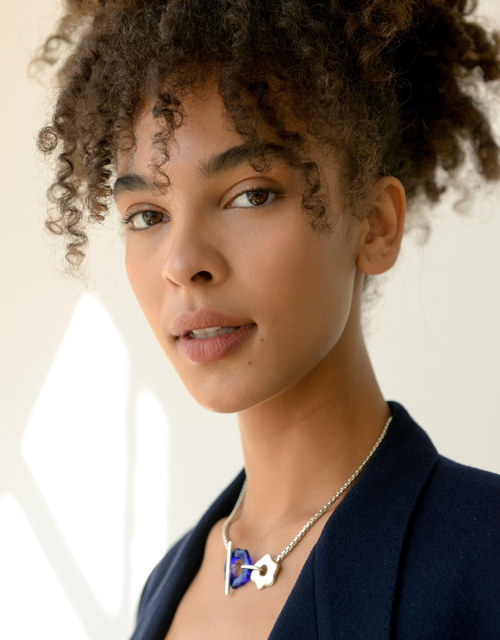 Elegant model wearing Avens Toggle Necklace, featuring abstract floral design in blue and white. Eco-friendly jewelry showcasing sustainable fashion and climate change awareness. Versatile toggle closure for multiple styling options. Handcrafted necklace from Floating Glacier Collection by CLED.