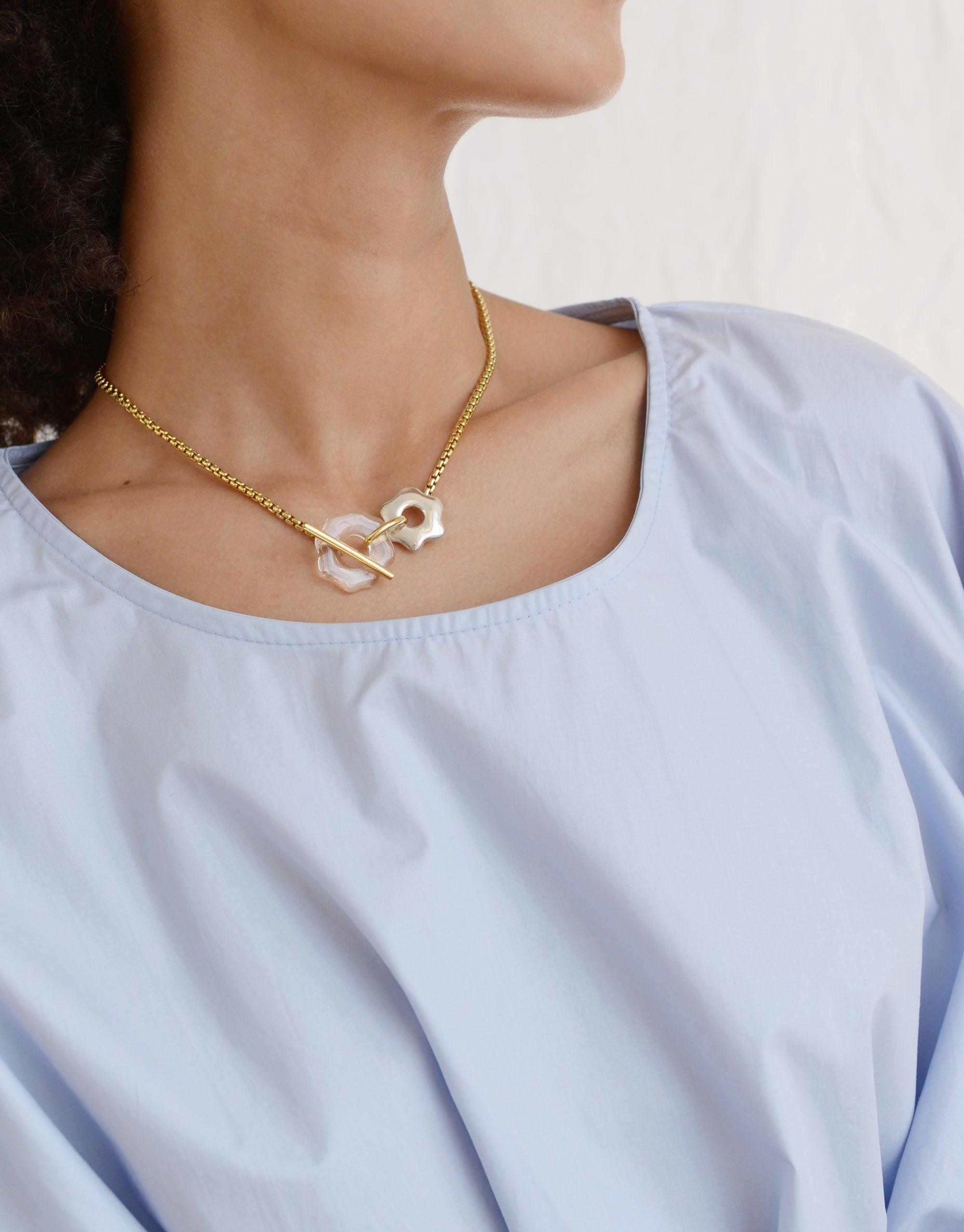 A person wearing a light blue top and the Avens Toggle Necklace, featuring a gold chain with a white abstract pendant inspired by the mountain avens flower. The necklace showcases a unique front toggle closure, part of the Floating Glacier Collection by CLED.