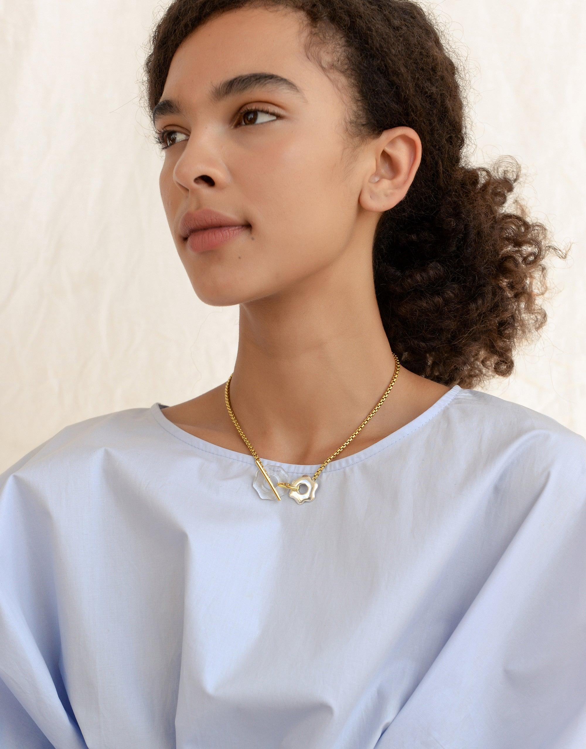 Elegant gold toggle necklace with abstract pendant inspired by Arctic avens flower, worn by model in light blue top. Versatile design with eco-friendly glass gem. Avens Toggle Necklace by CLED.