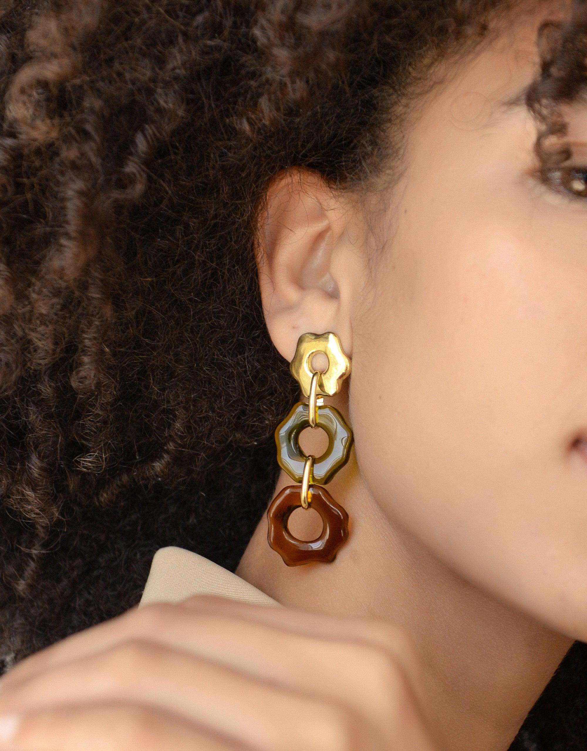 Close-up of Avens Earrings Trio featuring three colorful, organic floral-shaped charms in gold, blue, and brown, dangling from a model's ear. Handcrafted eco-friendly earrings showcasing unique design inspired by climate change awareness. CLED, Avens Earrings Trio.