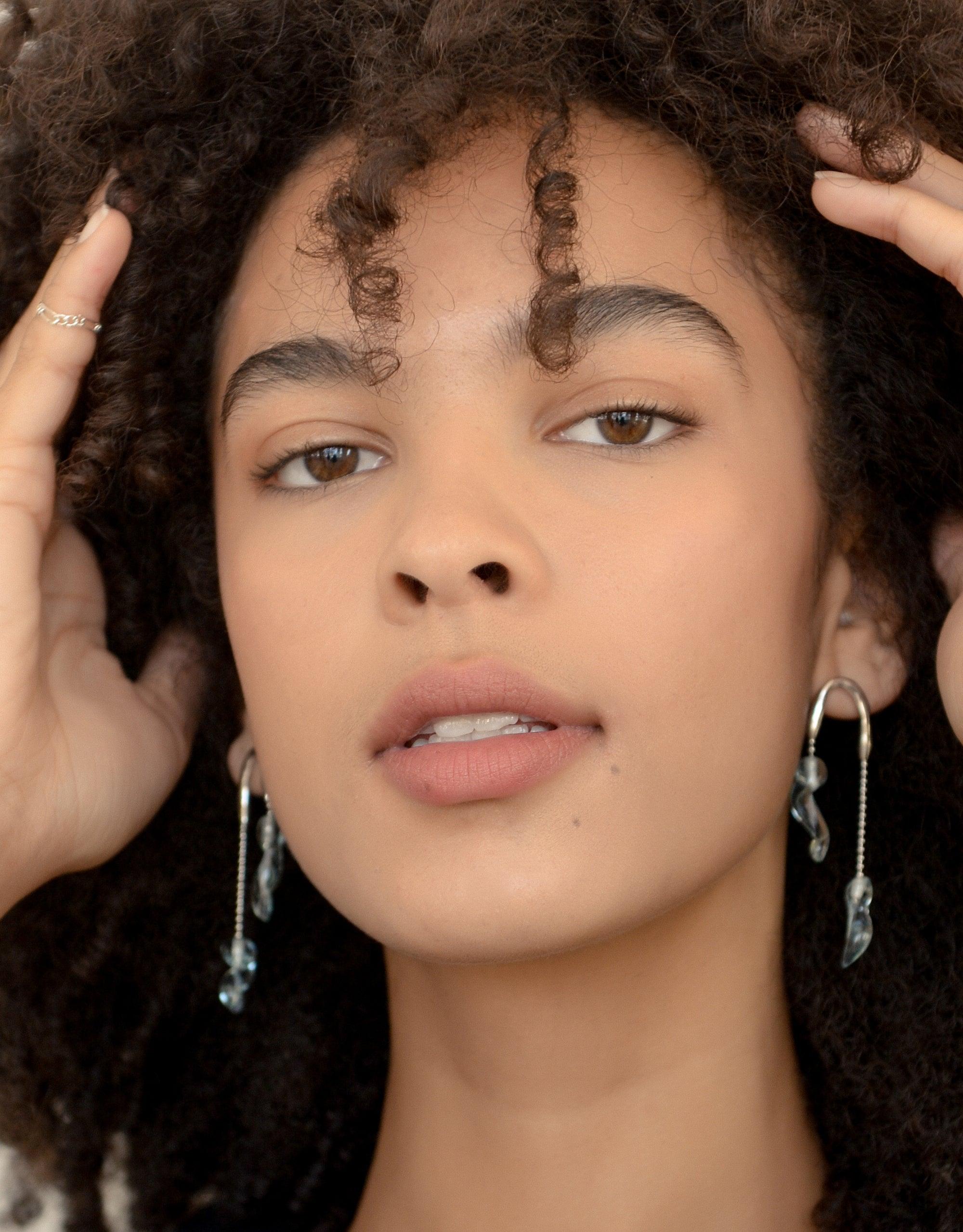 Close-up of a model wearing Crystal Drops Earrings, showcasing delicate asymmetrical icicle-inspired designs in sky blue. Hands gently touch curly hair, highlighting the earrings' elegant drop style. Eco-friendly jewelry by CLED.
